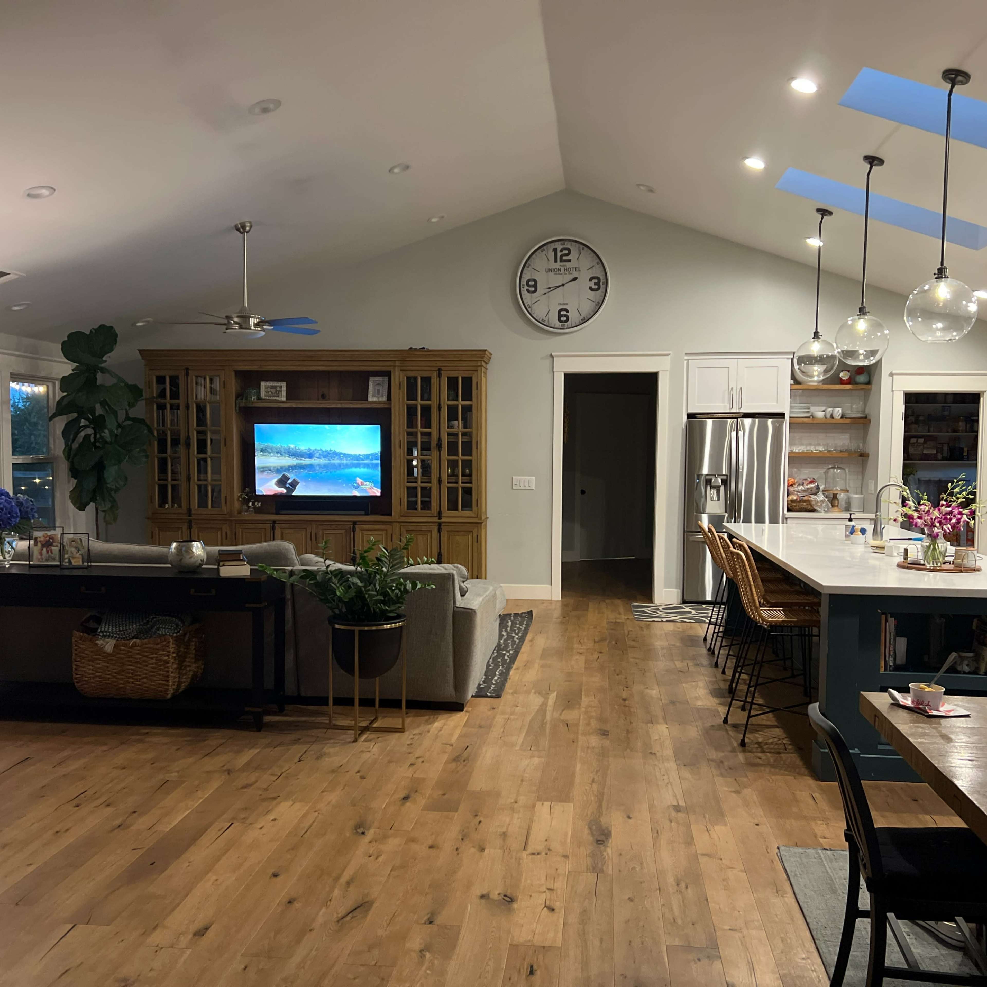 A modern living room and kitchen area with a large television, a dining table, and an open layout featuring wooden flooring and ample natural light.