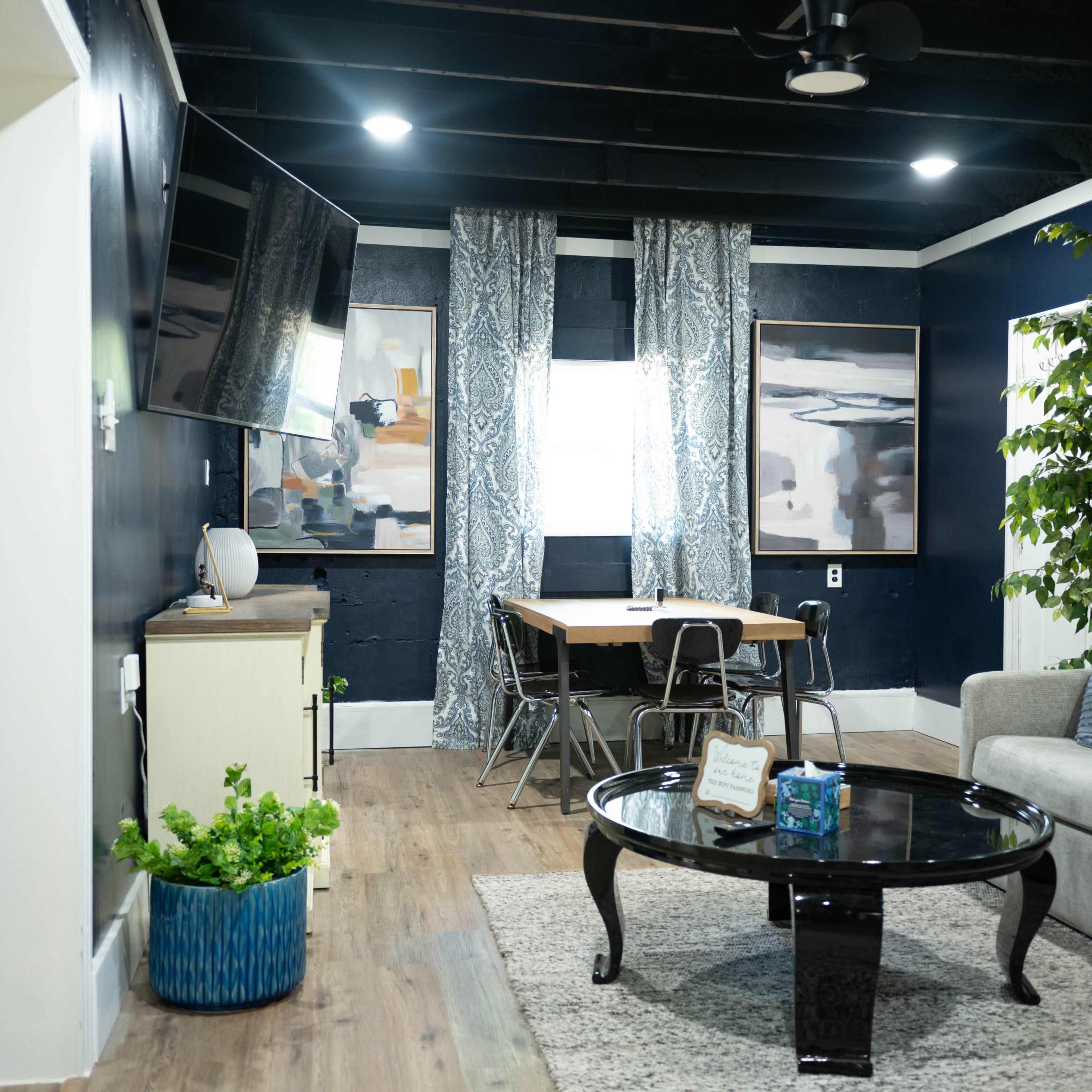A living room with a gray sofa, a circular black coffee table, a dining area with a wooden table, and large windows draped with patterned curtains.