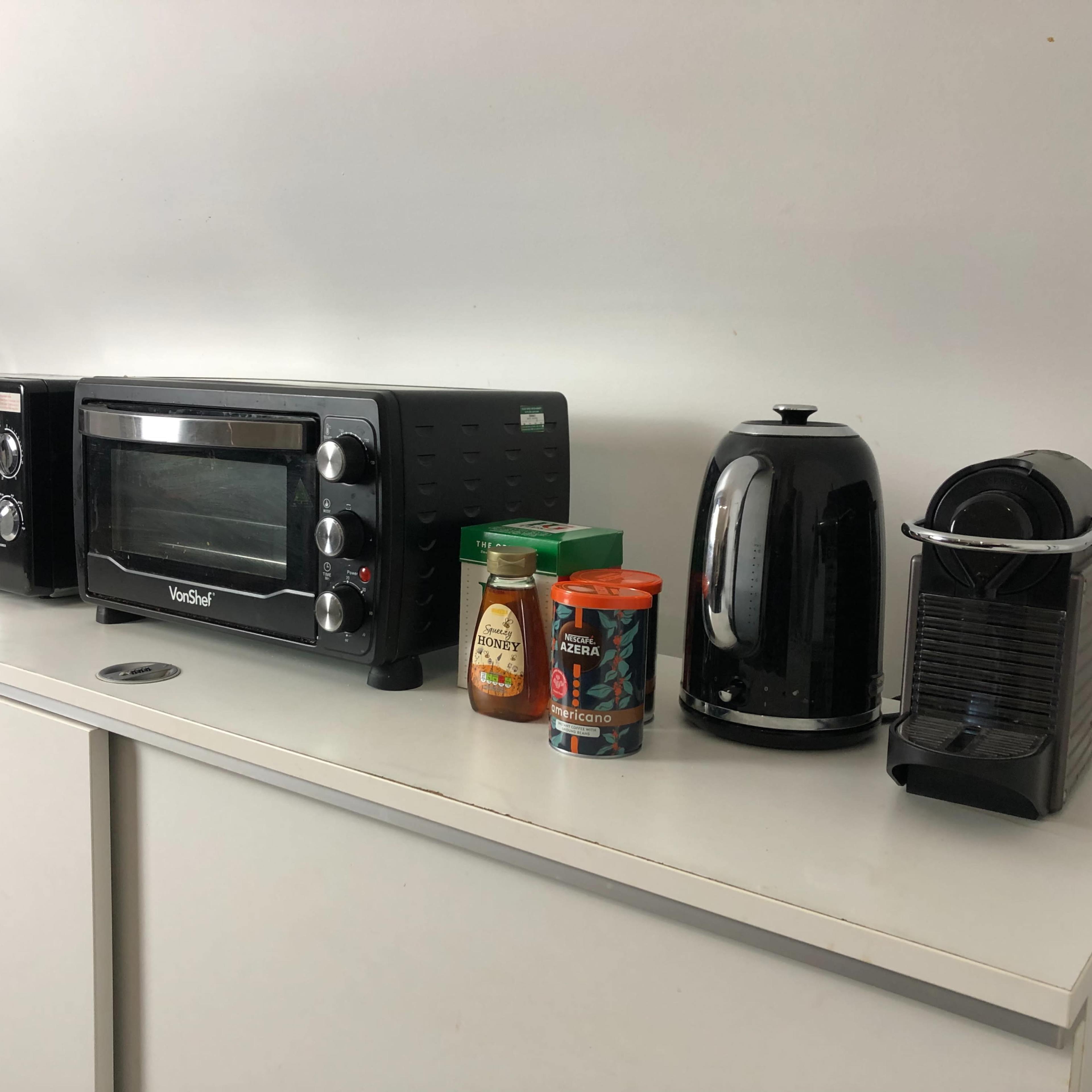 A kitchen countertop with a toaster oven, microwave, kettle, and coffee machine, accompanied by jars of honey and coffee.