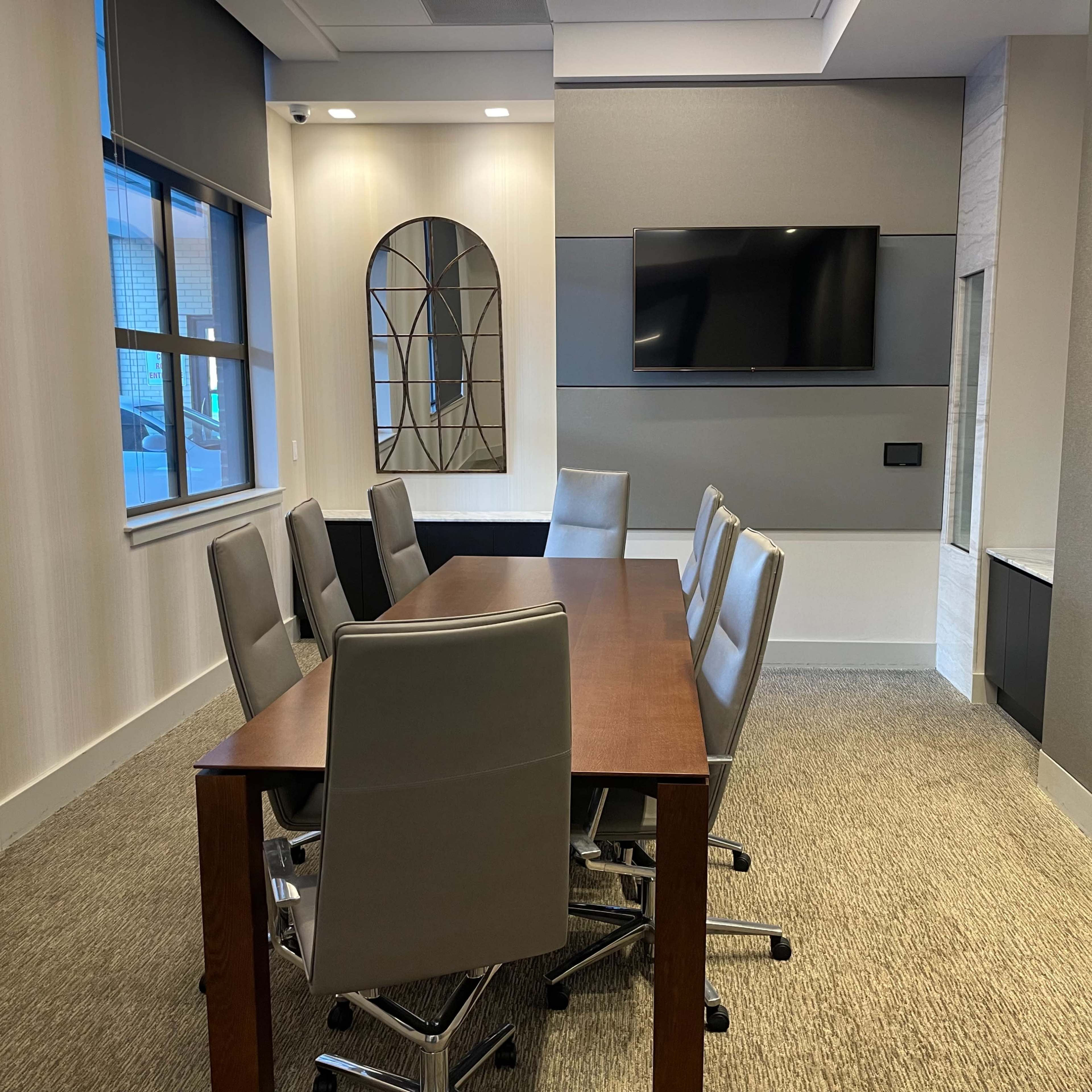 A meeting room features a wooden table surrounded by six ergonomic chairs, a wall-mounted television, and large windows.