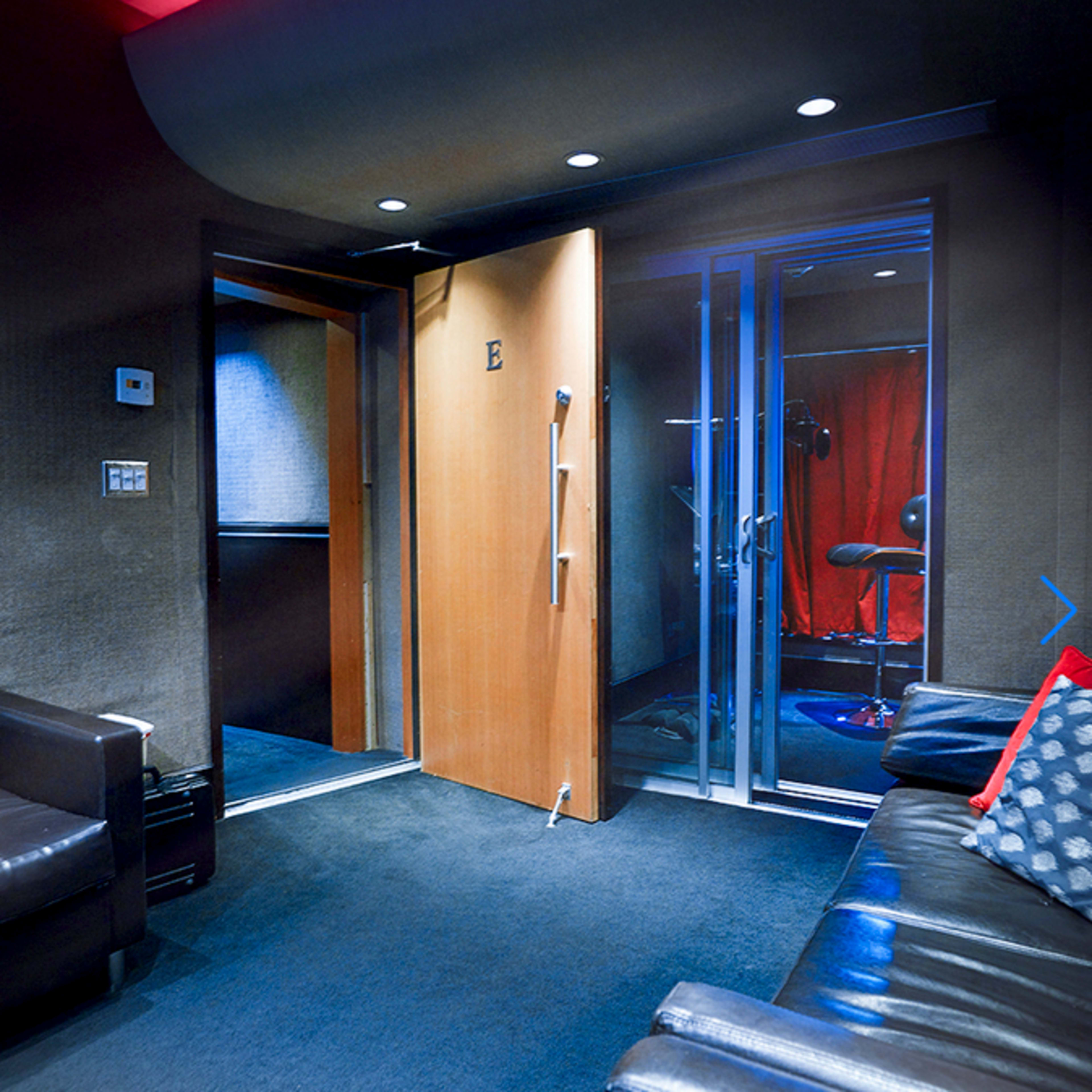 A modern room with dark walls, a partially open wooden door leading to a hallway, and glass doors that likely open to a soundproof booth.