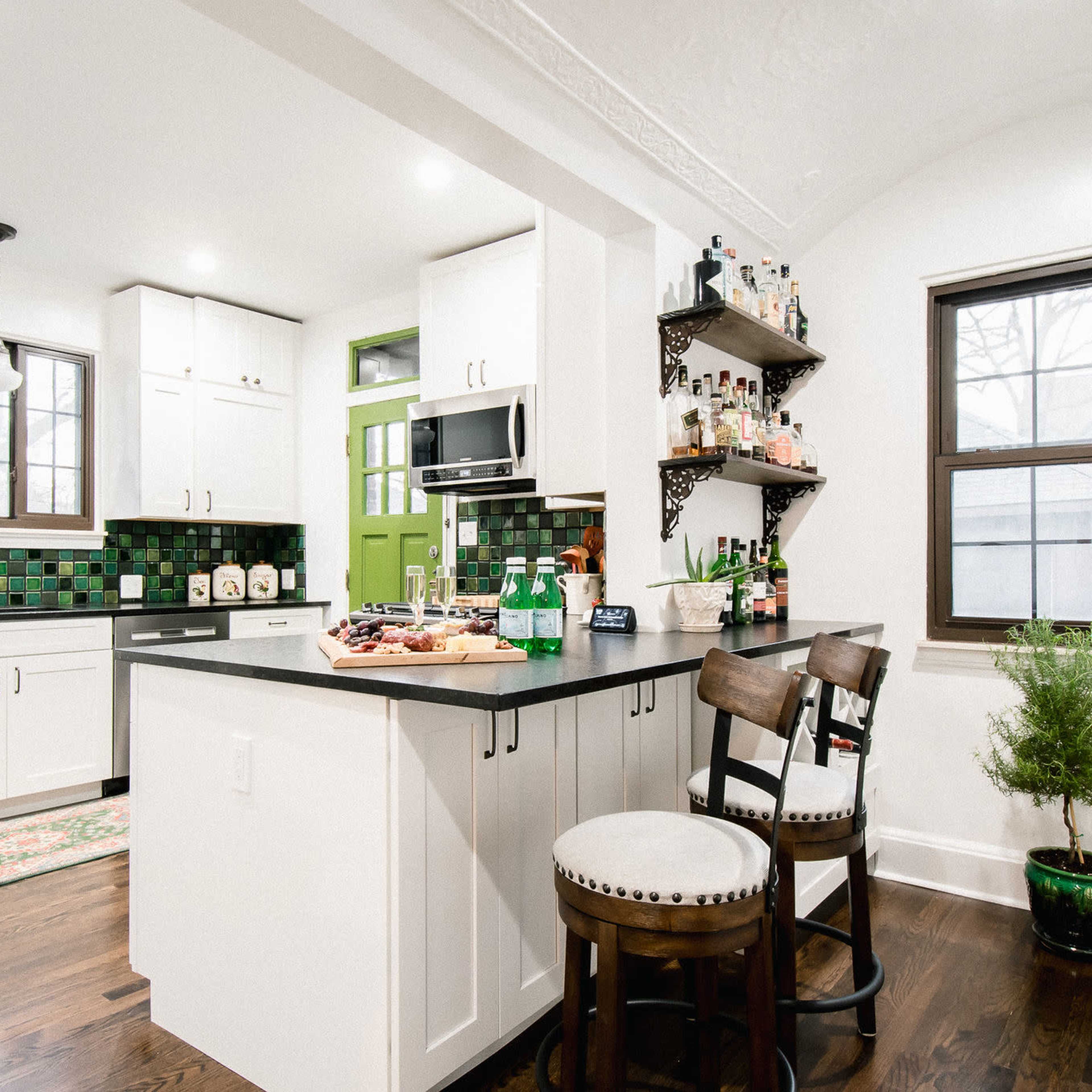 A modern kitchen featuring white cabinets, green accent walls, a dark countertop, and a small dining area with two chairs and a bar shelf displaying various bottles.