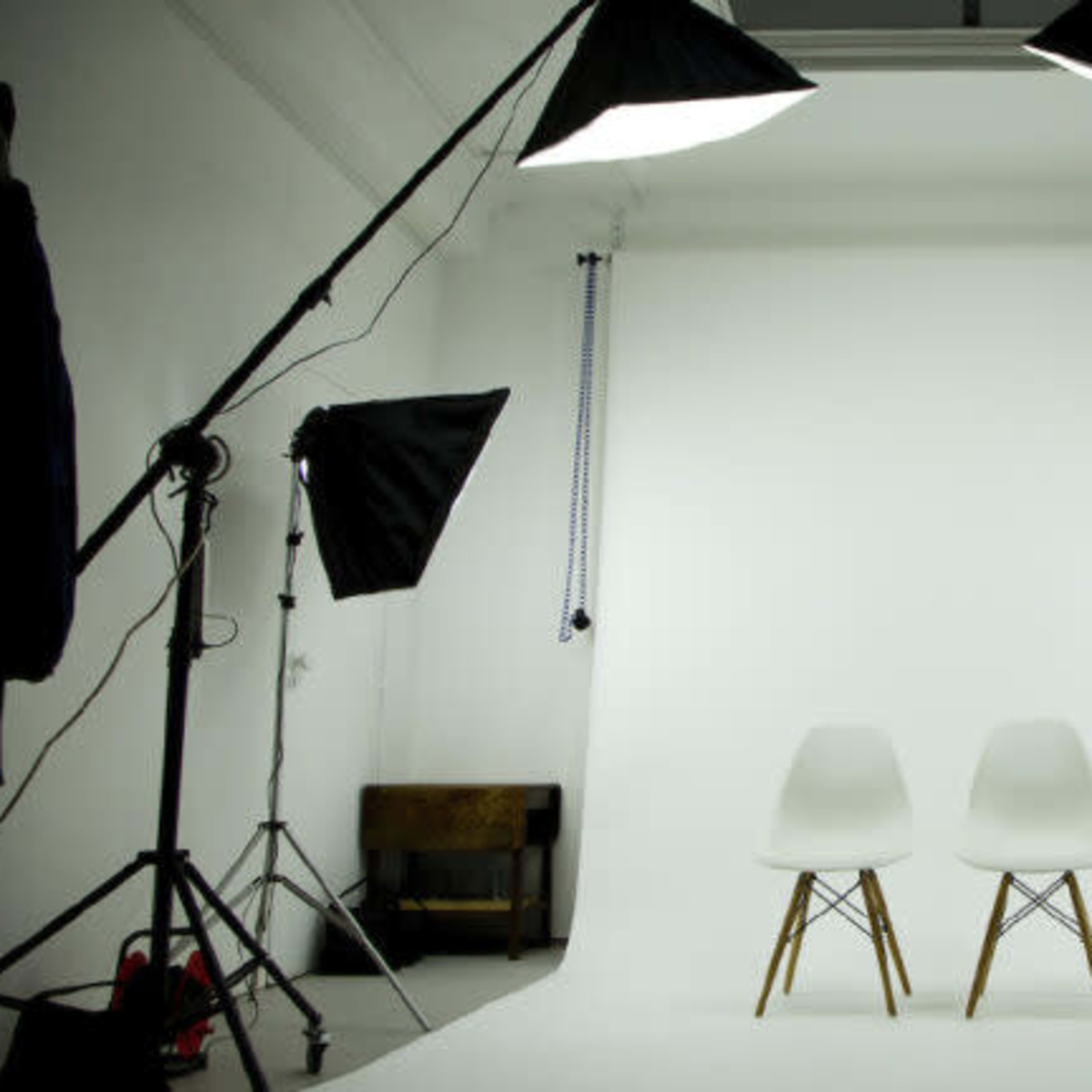 A photography studio equipped with two white chairs centered on a bright backdrop, surrounded by multiple lighting setups.