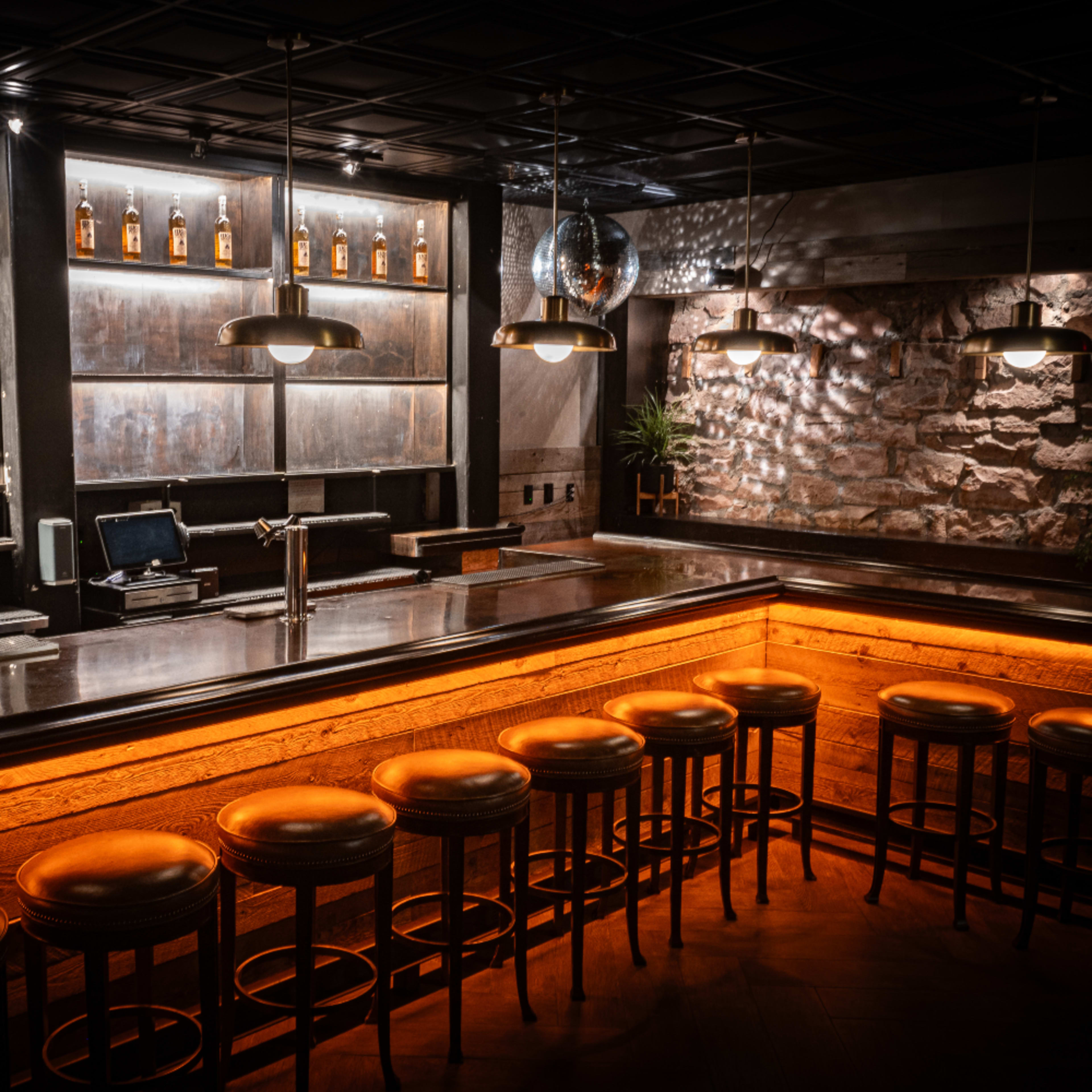 A dimly lit bar with a polished countertop, wooden stools, and shelves displaying bottles of liquor.