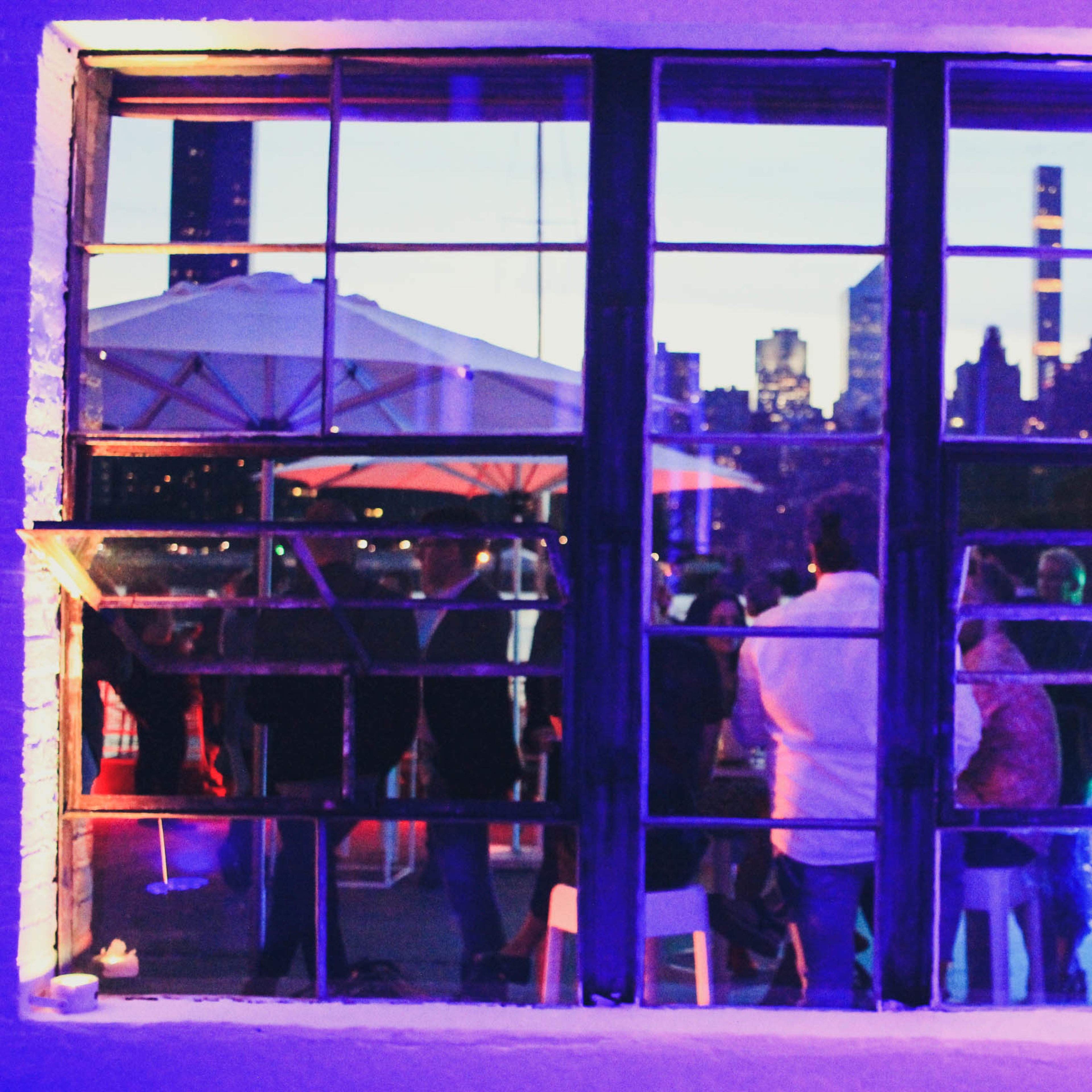 People gathered inside a venue with large windows overlooking a city skyline at dusk.