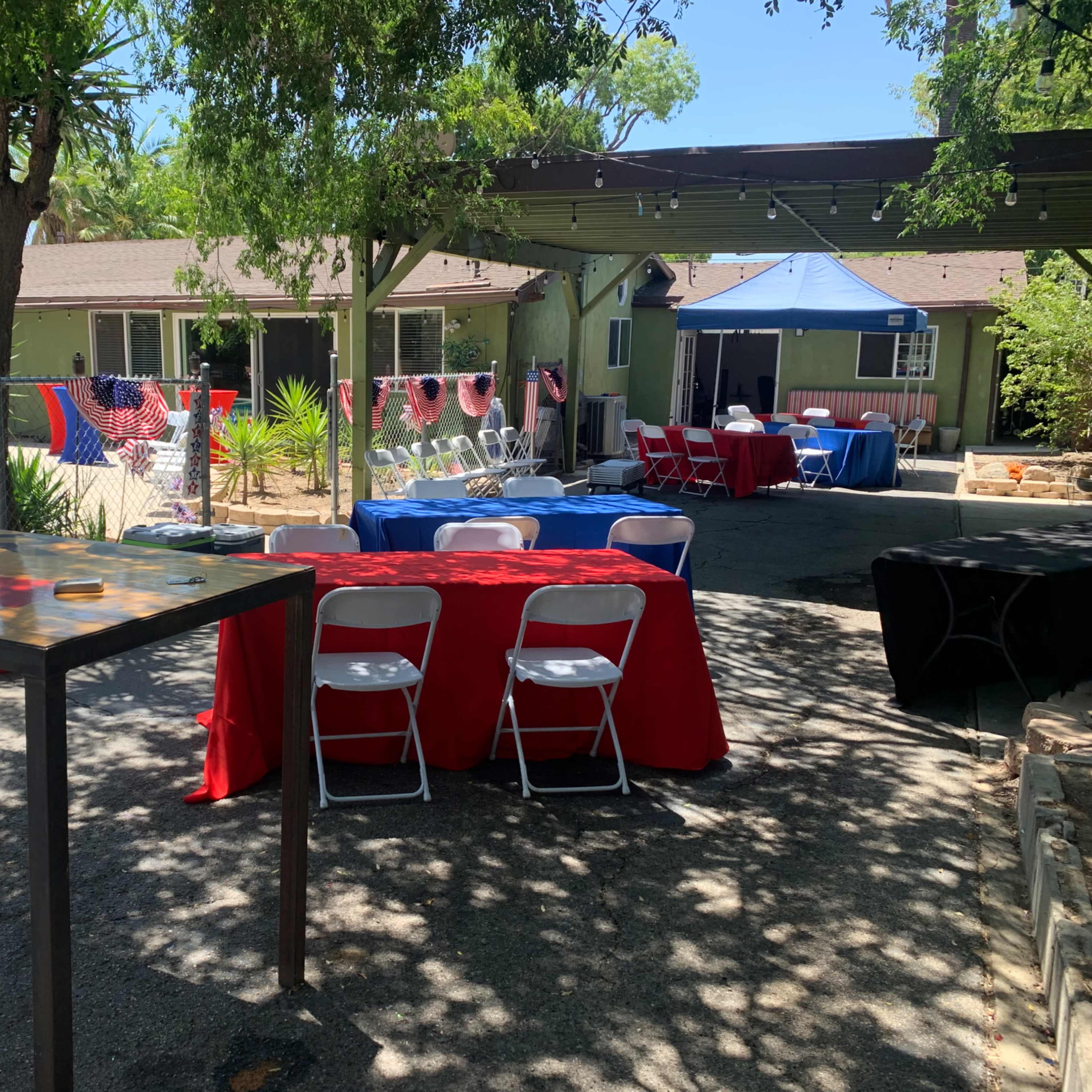 An outdoor event space featuring several tables with red and blue tablecloths, seating, and decorations near a swimming pool and a large tent.