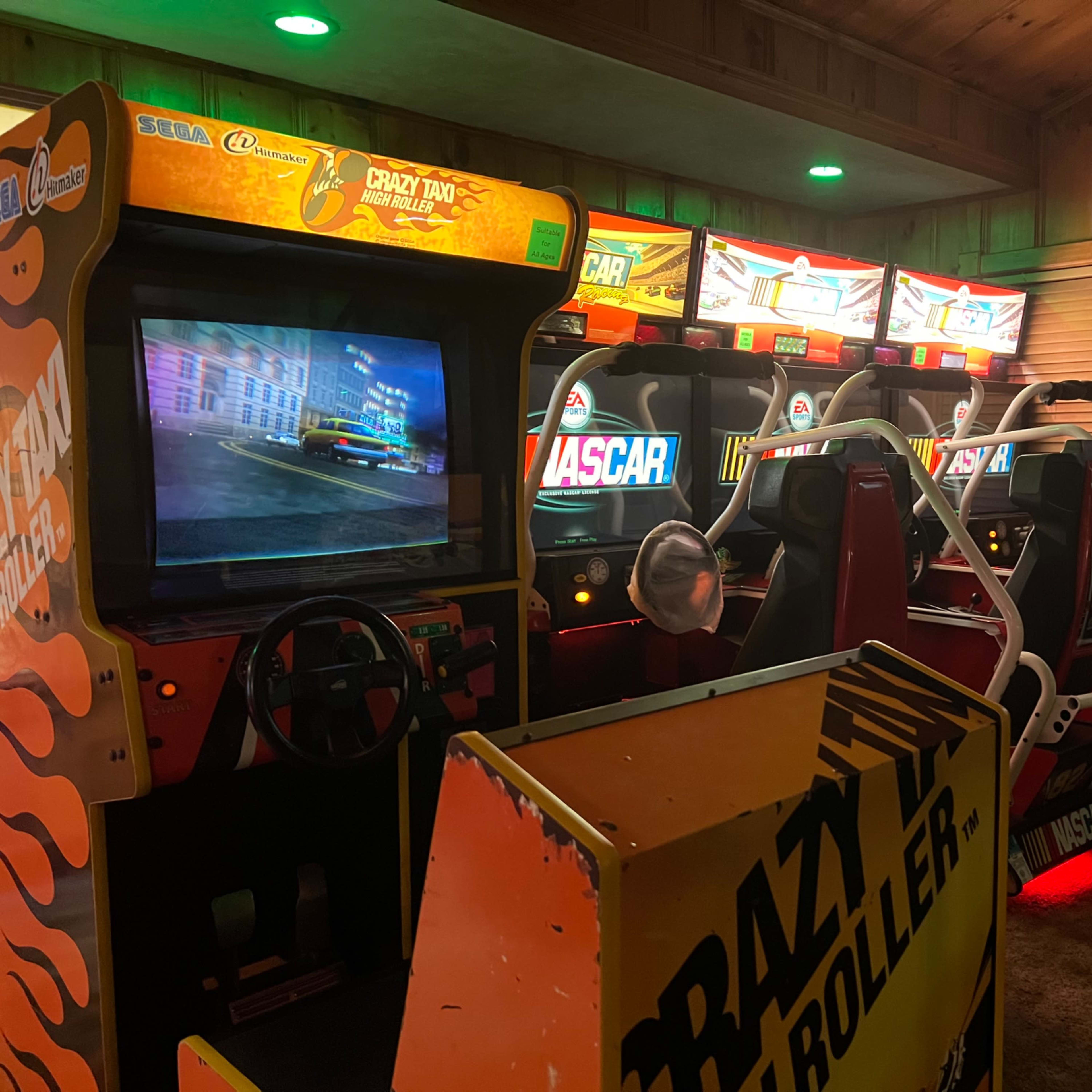 A row of classic arcade racing games in a dimly lit room, with a child seated at one of the machines.