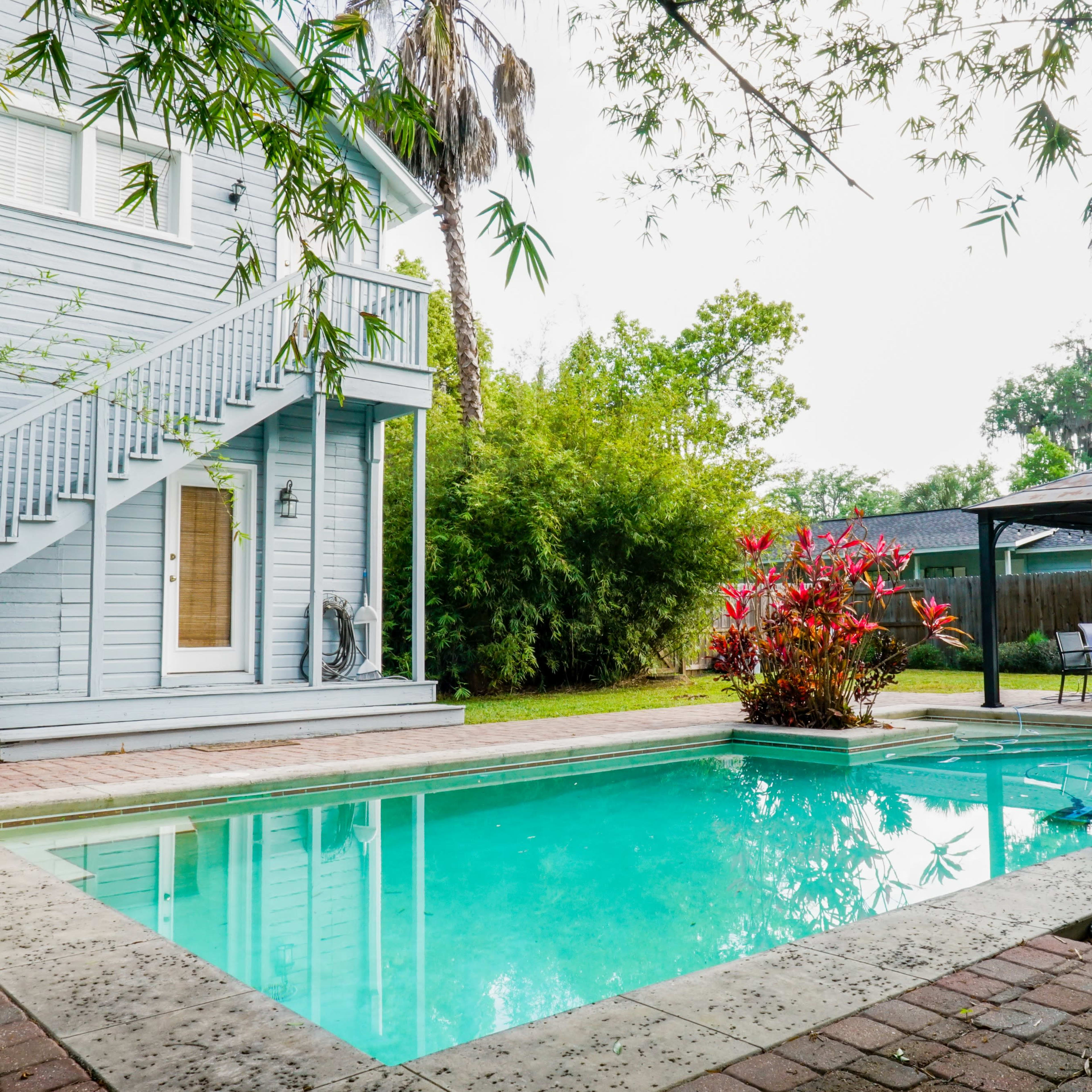 A clear swimming pool is surrounded by a stone patio and lush greenery next to a two-story blue house.