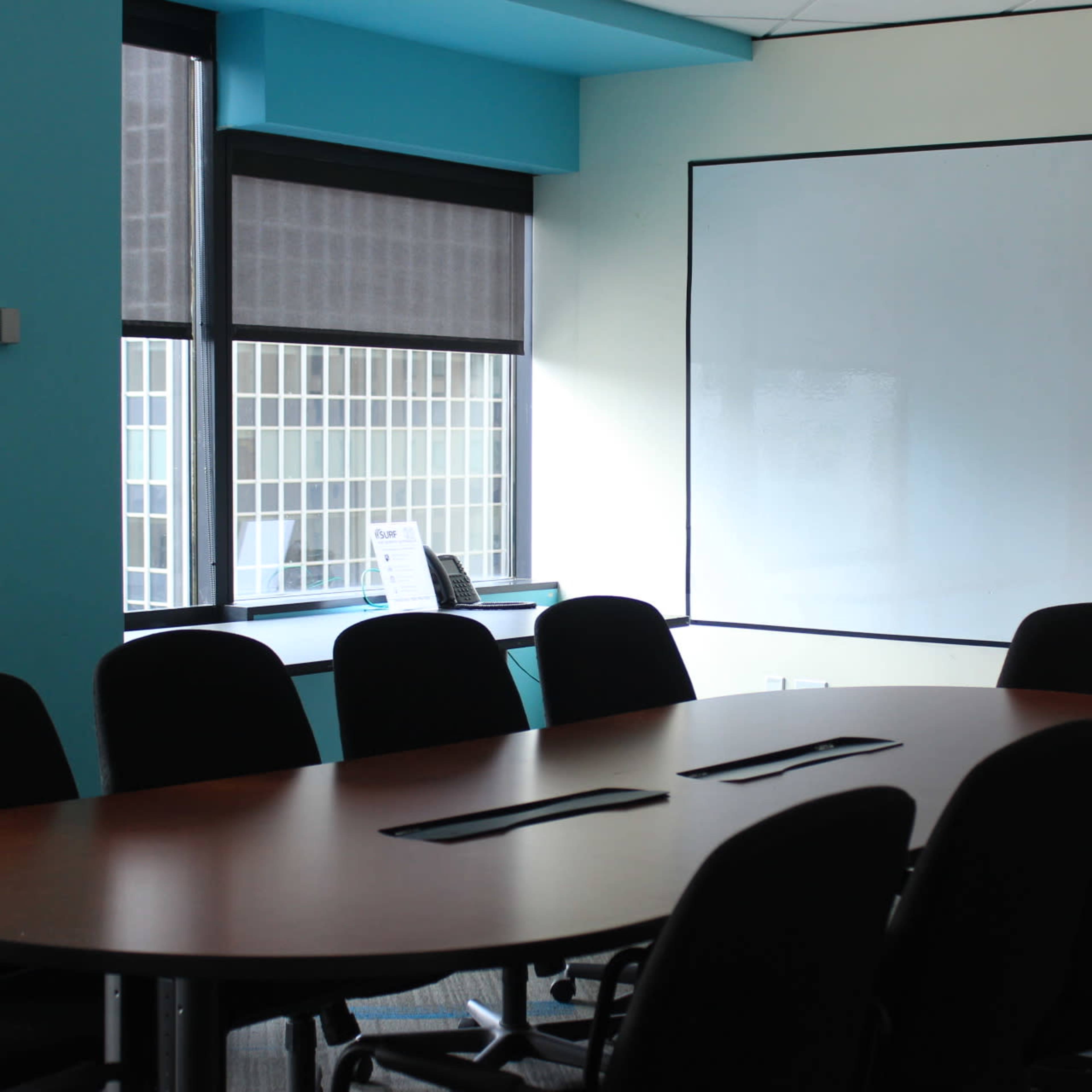 A conference room with a large wooden table surrounded by black chairs, and a whiteboard on one wall next to a window.