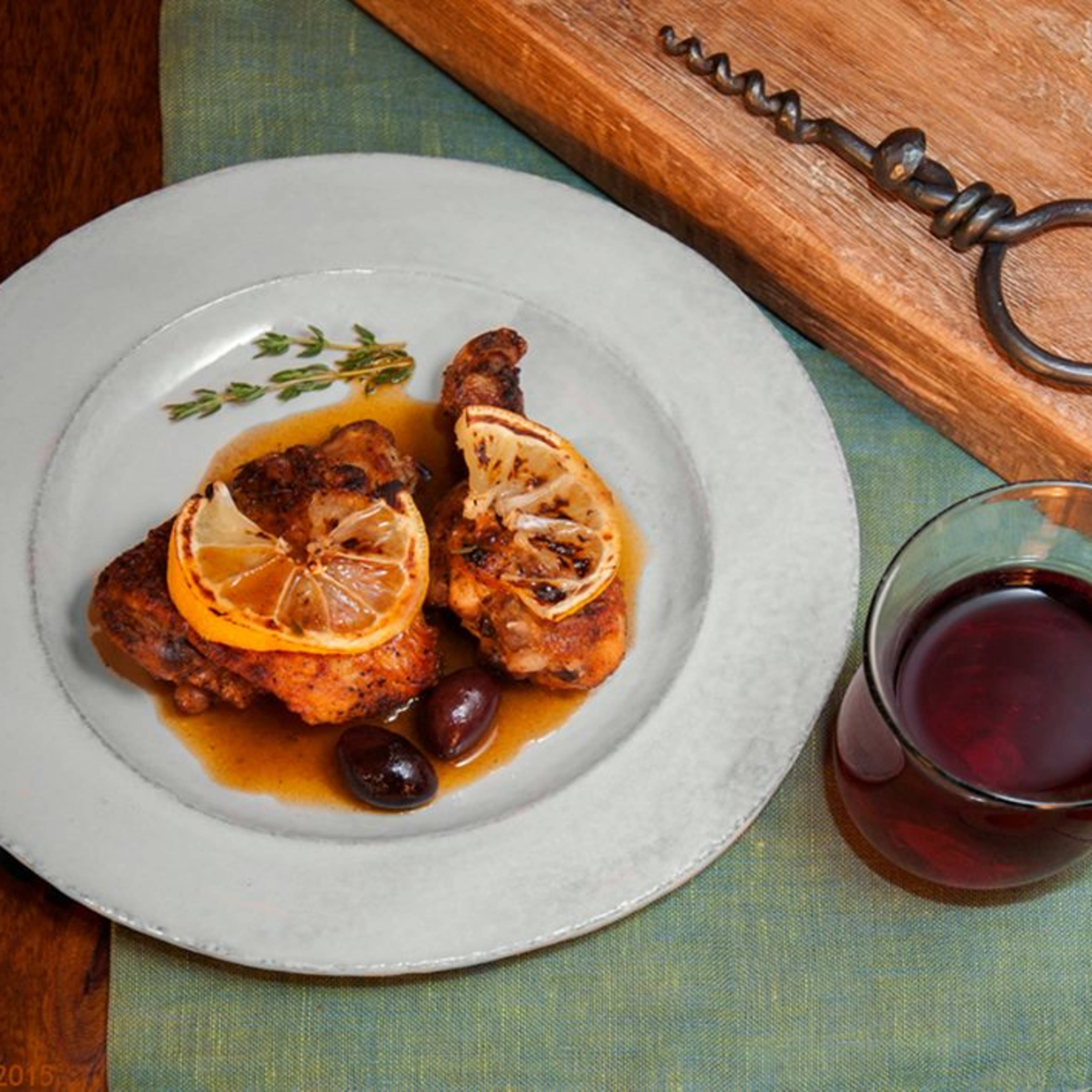 A plate of chicken drumsticks garnished with lemon slices and olives, accompanied by a glass of red wine on a wooden table.