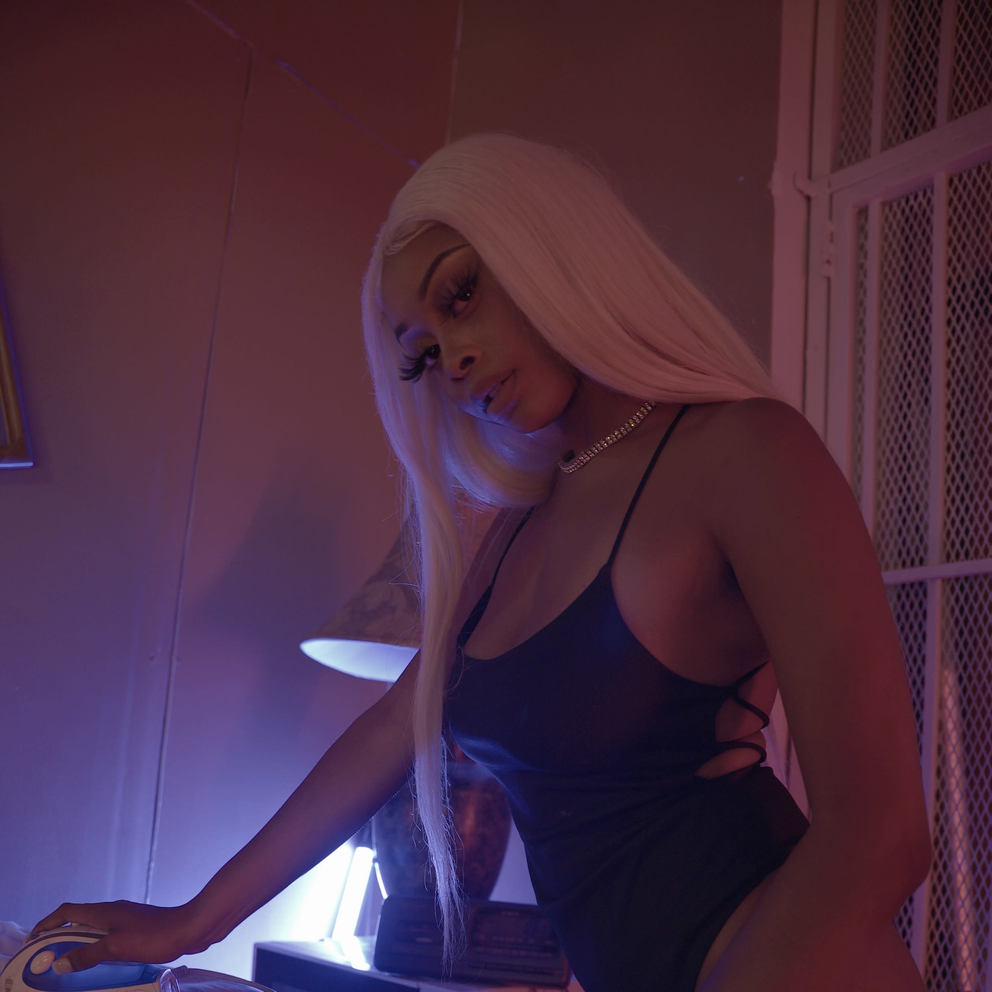 A person in a black outfit is ironing clothes while surrounded by money on an ironing board in a dimly lit room.