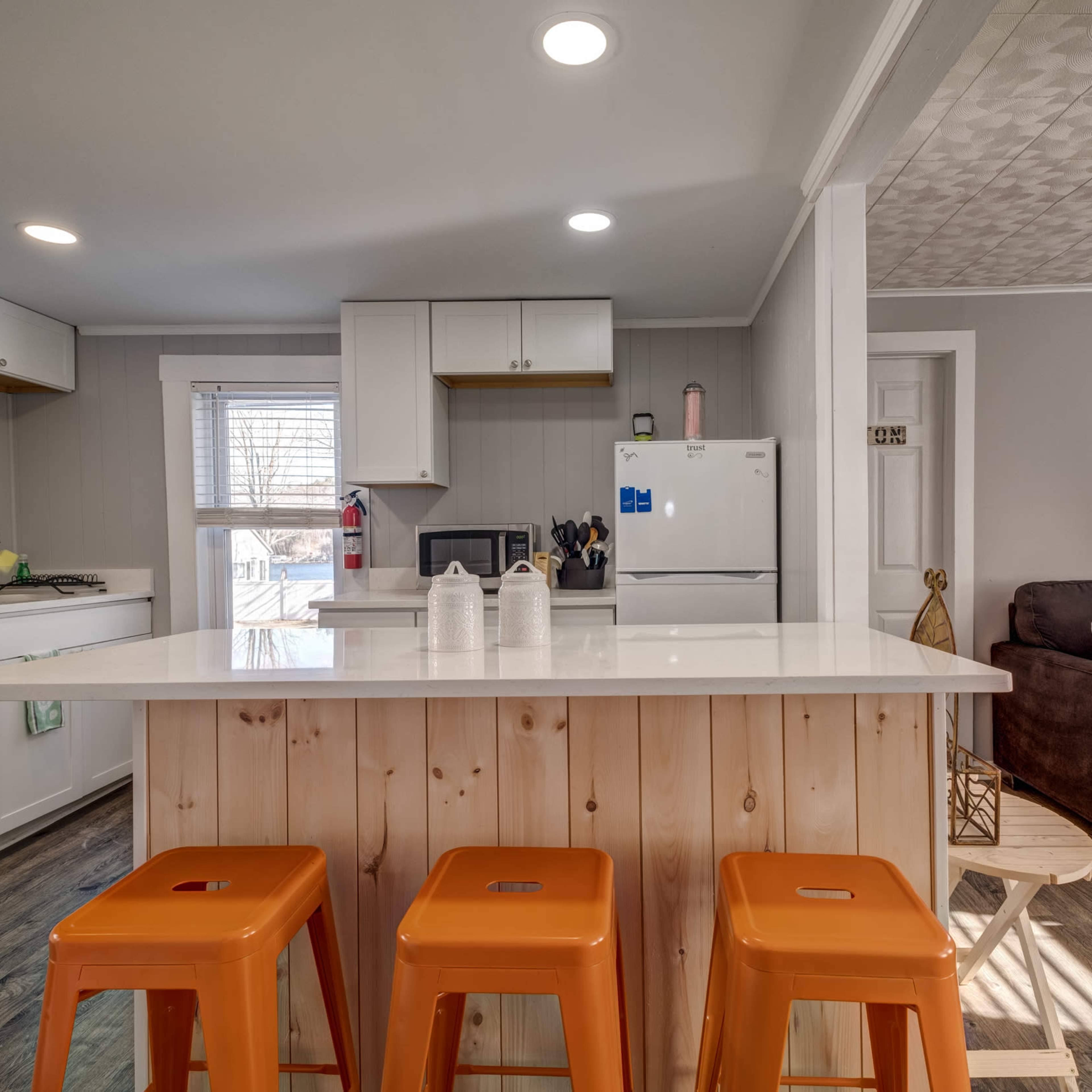 A modern kitchen with white cabinets, a central island with three orange stools, and a living area featuring a brown couch in the background.