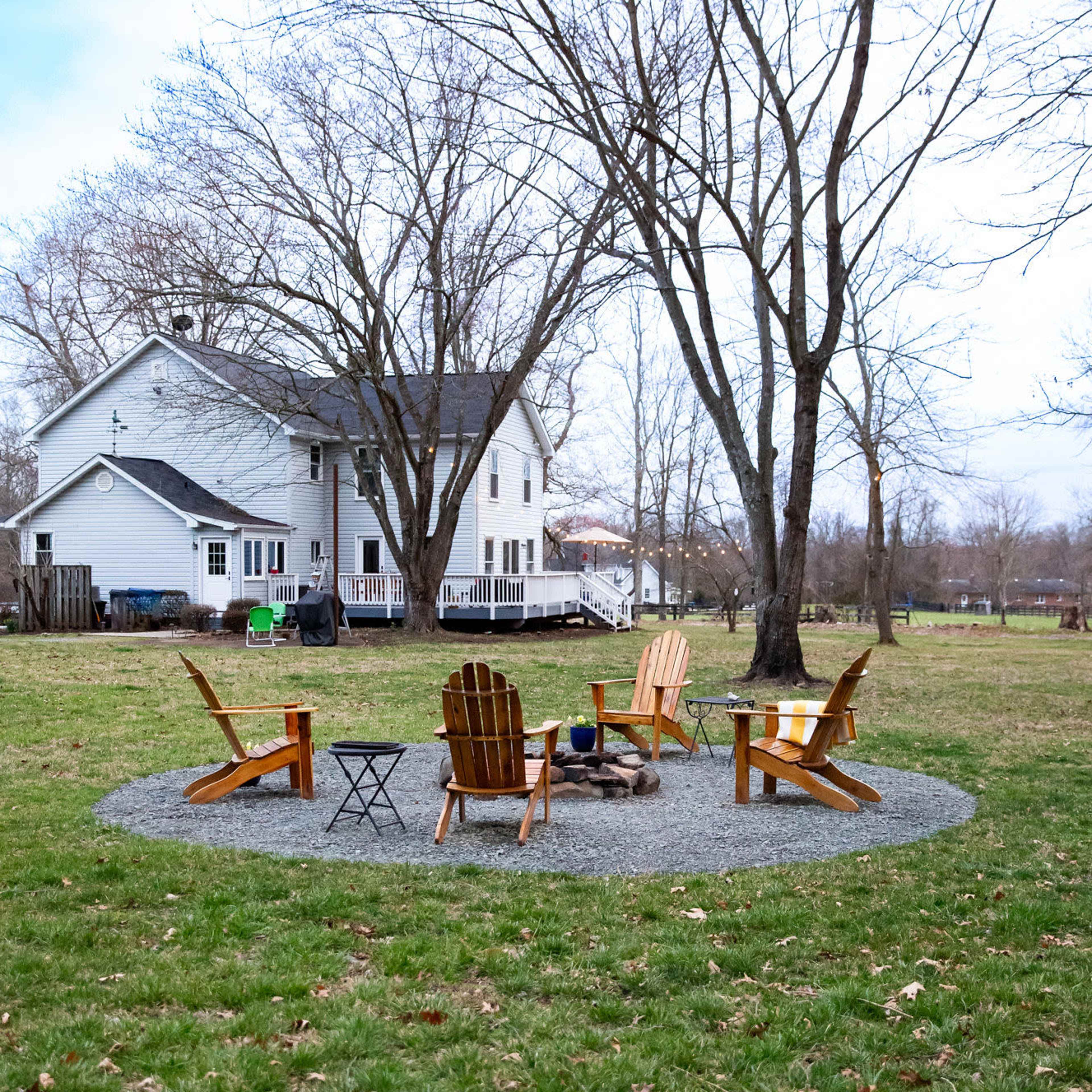 A circular gravel area with four wooden Adirondack chairs surrounding a fire pit is situated in a grassy yard, with a white house and bare trees in the background.