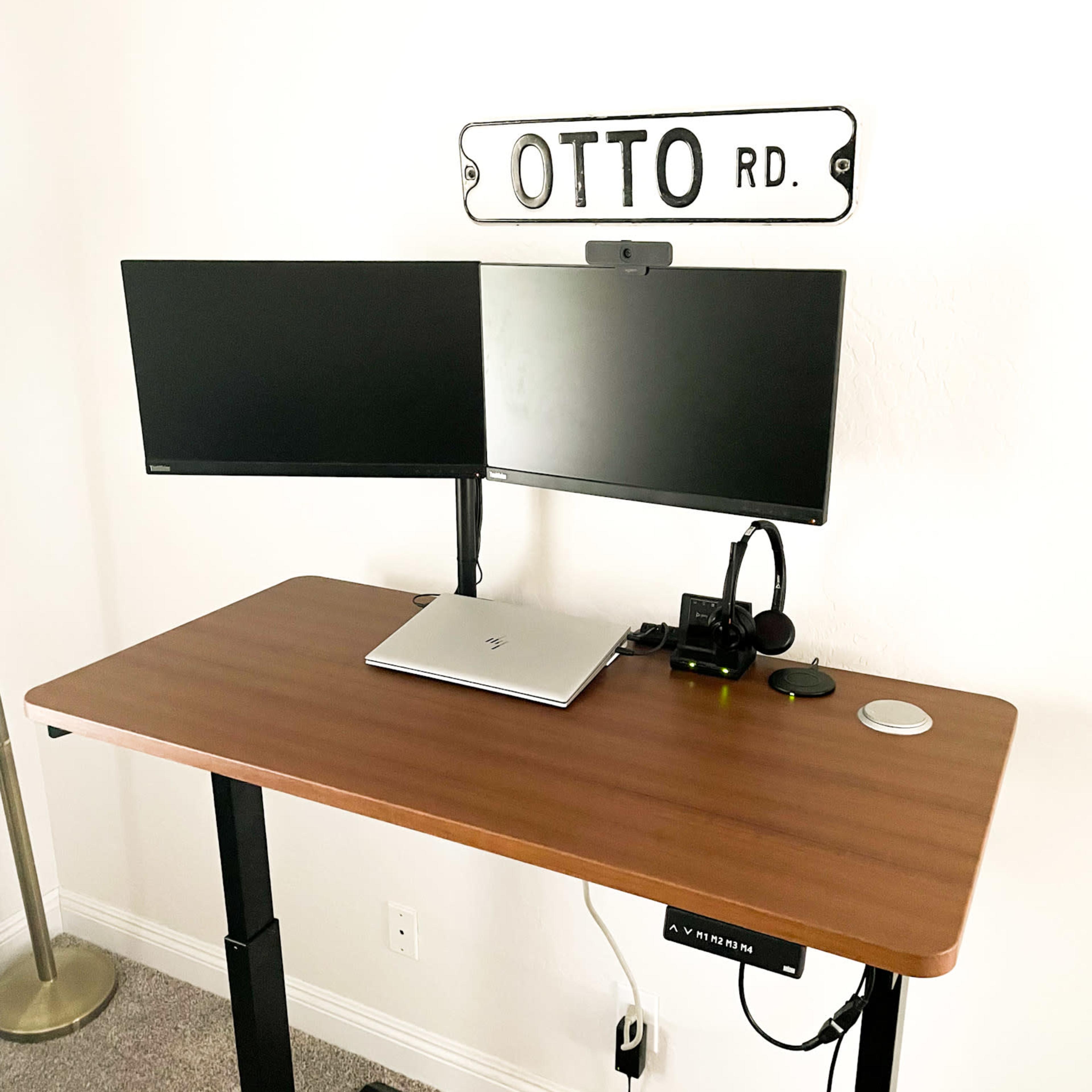 A standing desk with two monitors, a laptop, and a webcam is positioned near a wall with a sign that reads "OTTO RD."