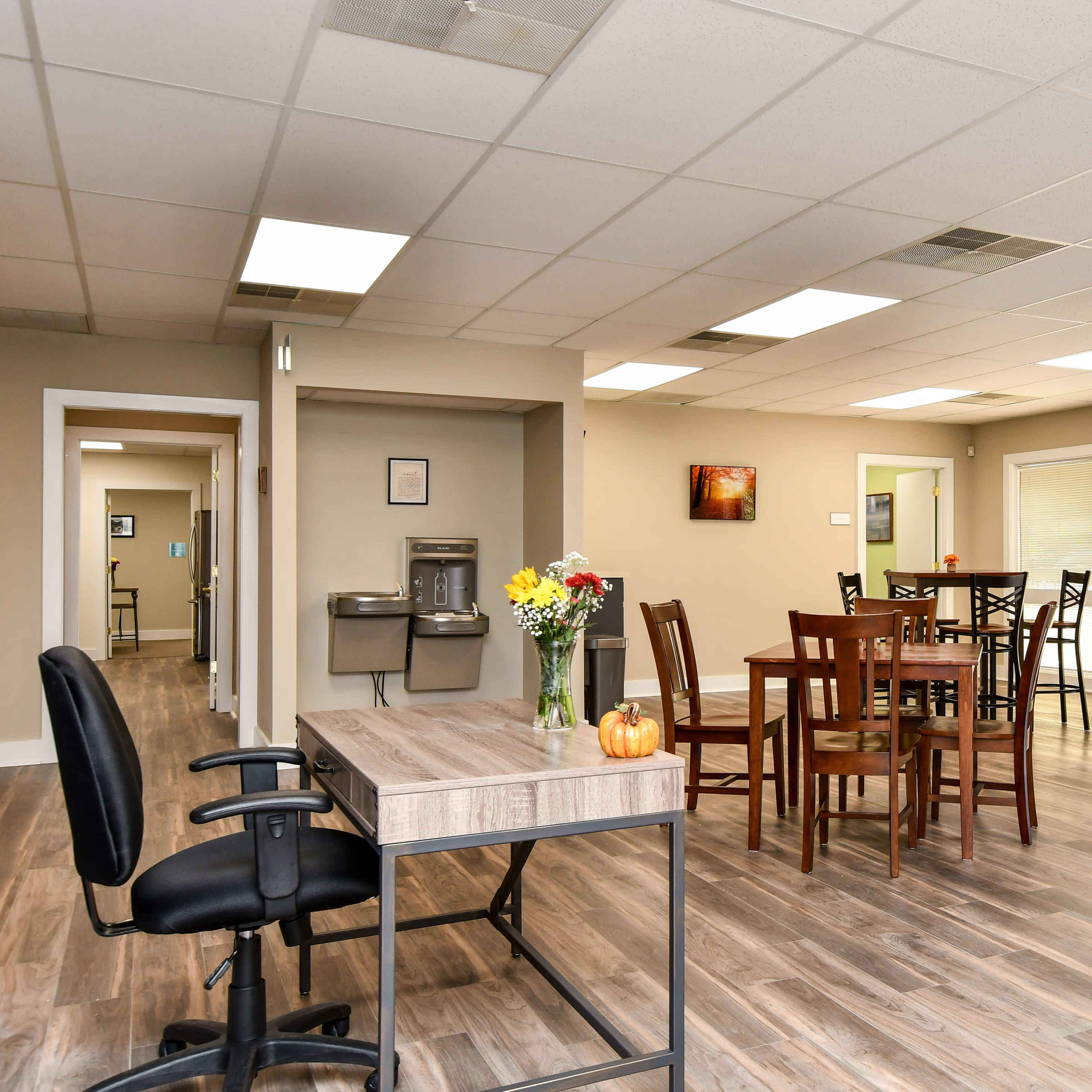 An indoor space featuring tables and chairs, a wooden desk with a swivel chair, a drinking fountain, and large windows allowing natural light.