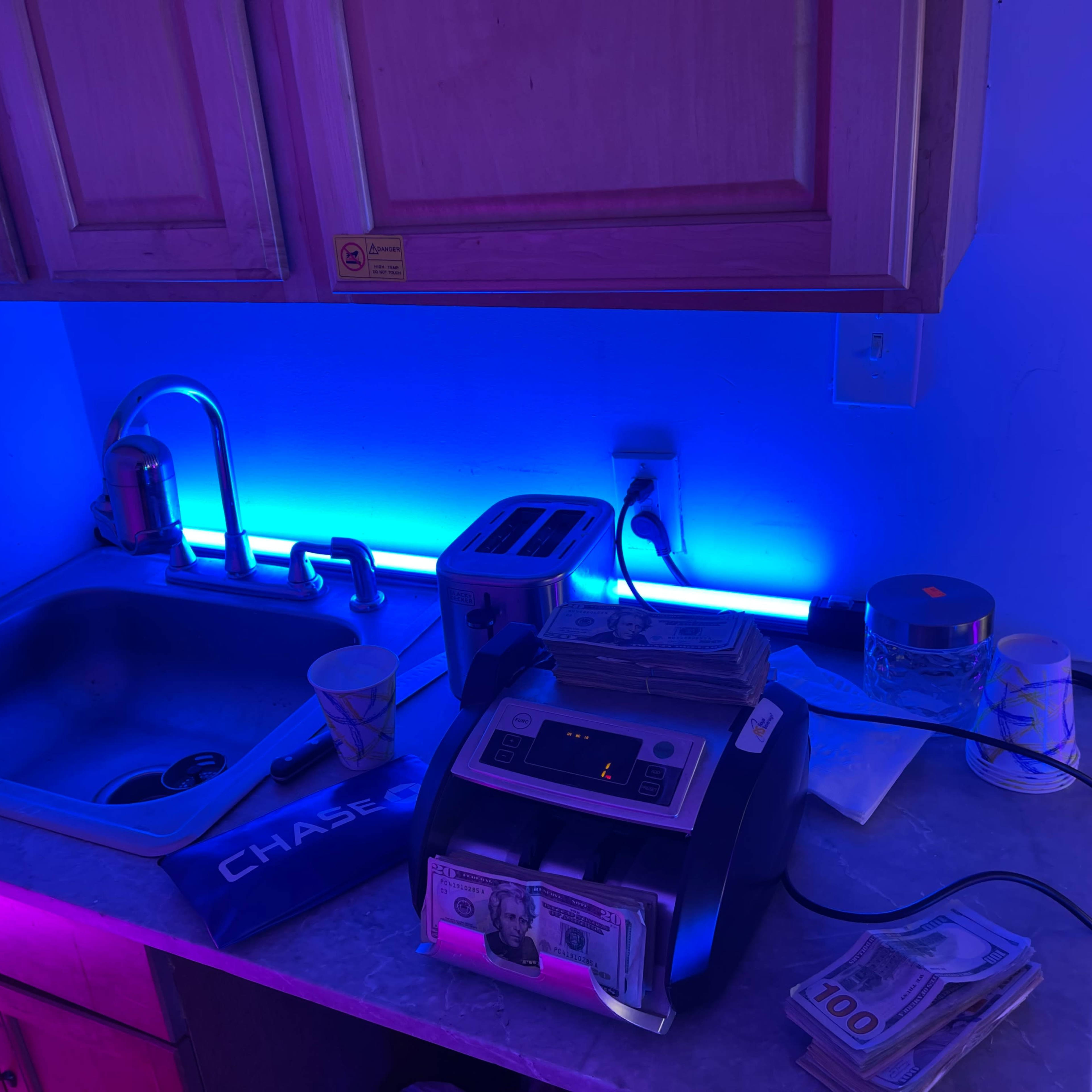 A countertop features a money counting machine, stacks of cash, a toaster, and a sink, illuminated by blue LED lights.