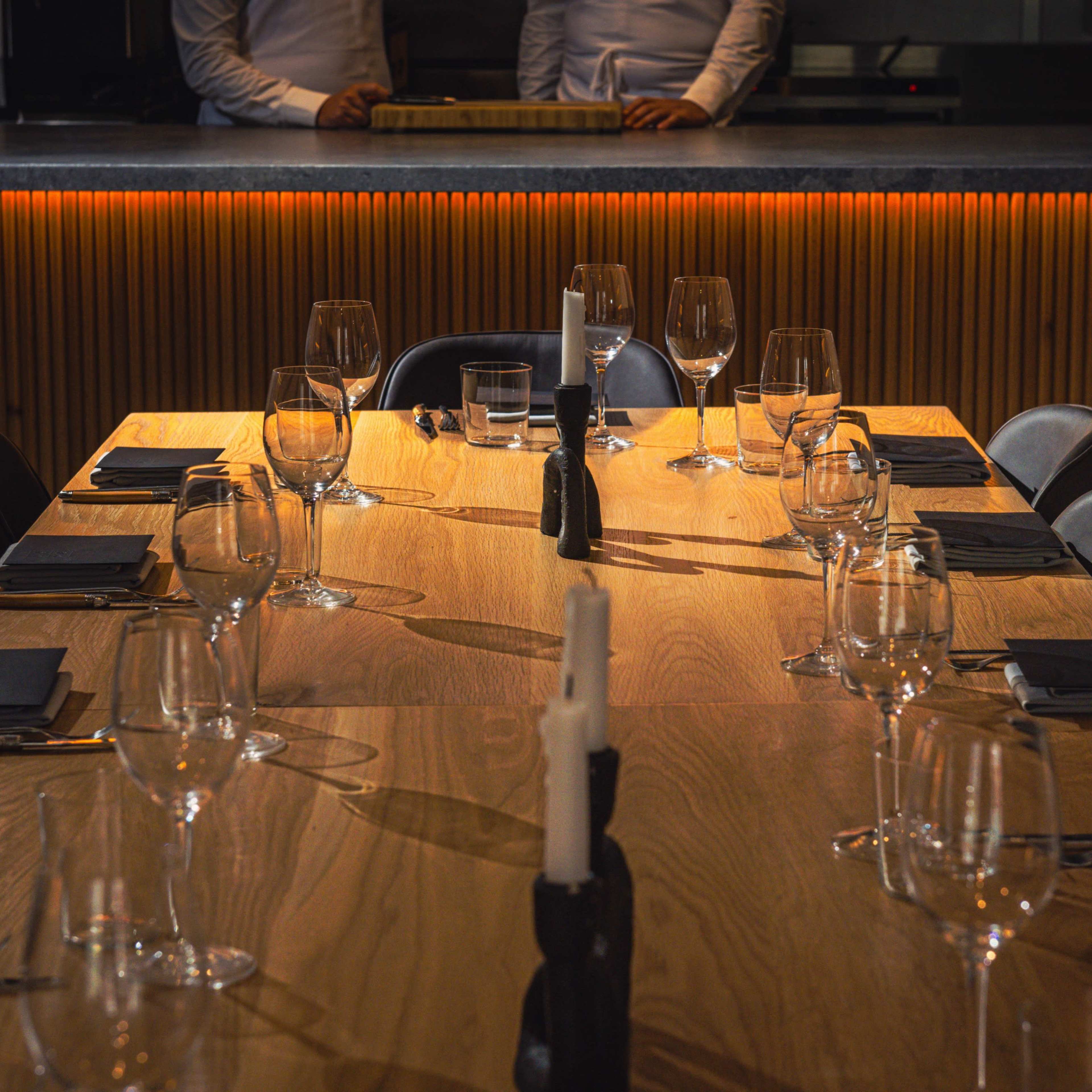 A large wooden dining table is set with glasses and black napkins, illuminated by warm light from the background.