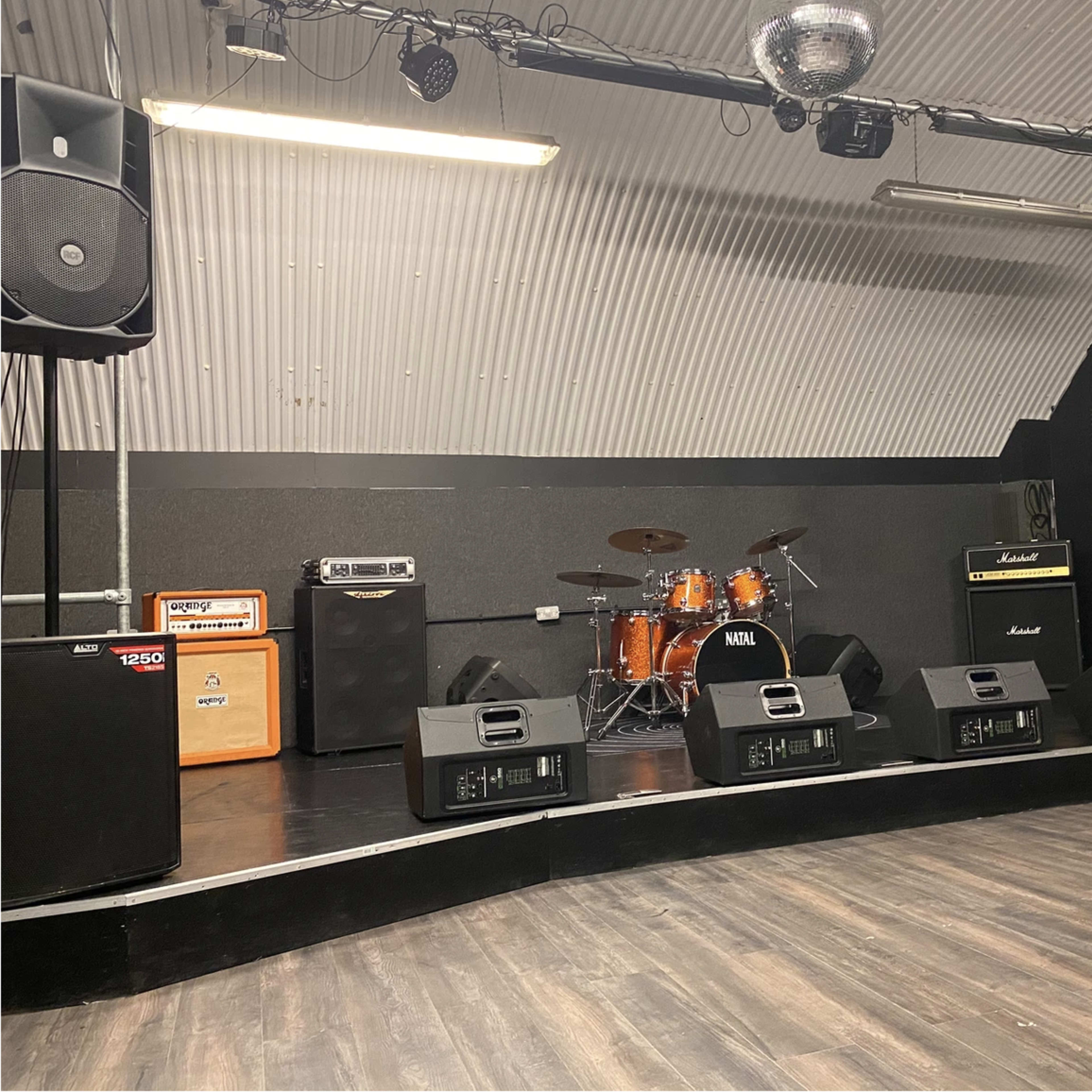 A stage setup with several amplifiers, drums, and speakers in a curved room with wooden flooring and a disco ball overhead.