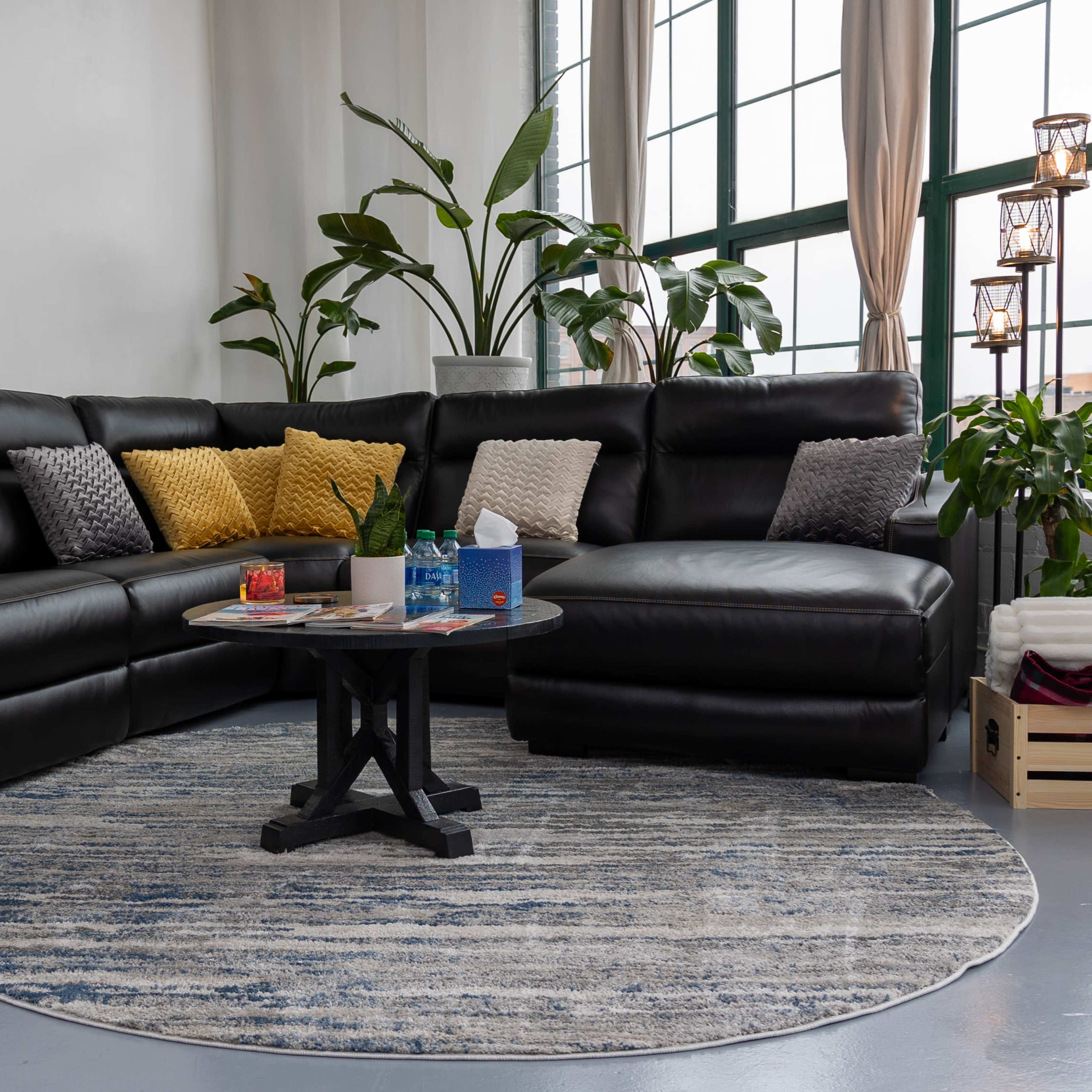A spacious living area features a large black leather sectional couch, a round area rug, a coffee table with several items on it, and several potted plants by the window.