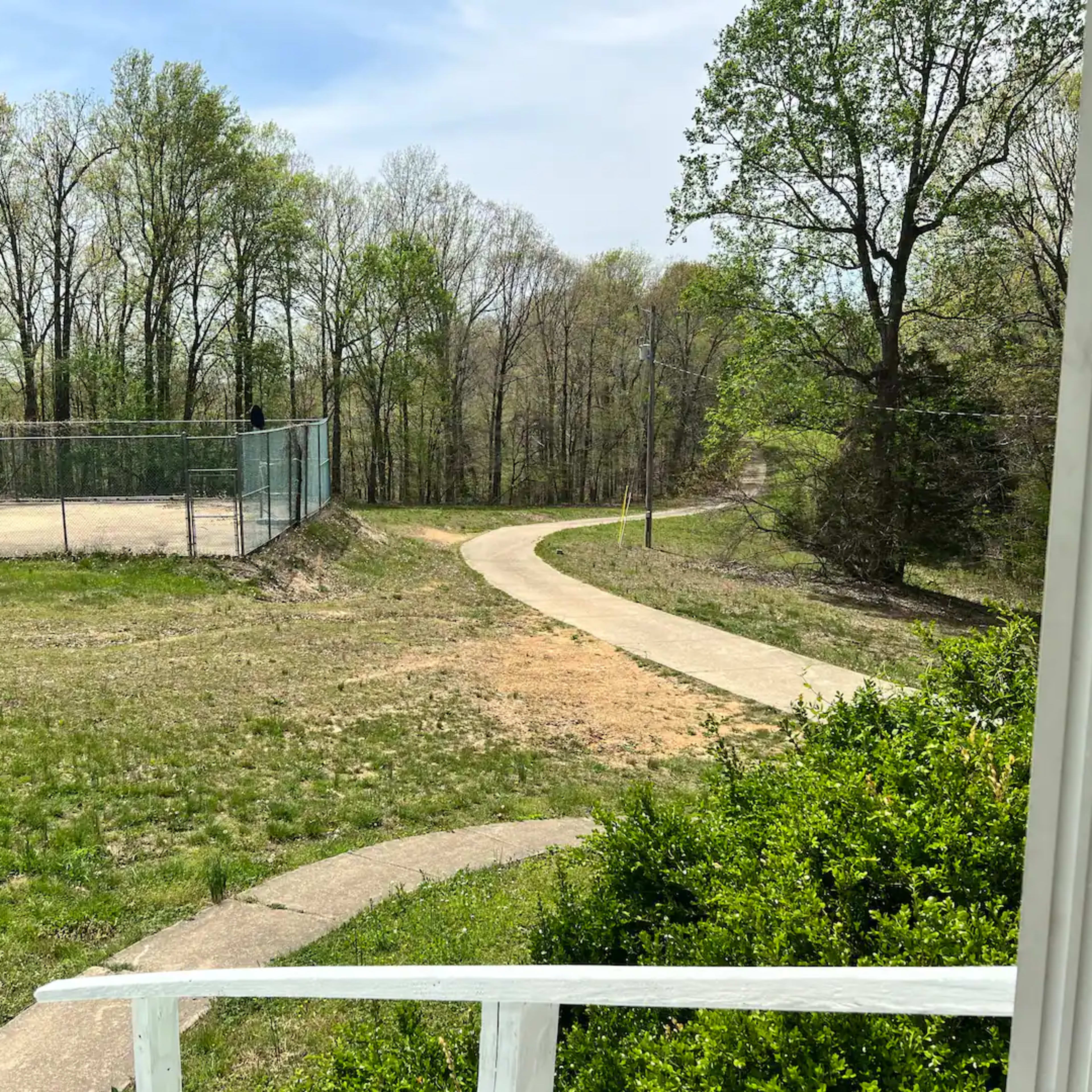 A view of a winding concrete path leading through a grassy area toward a baseball field surrounded by trees.