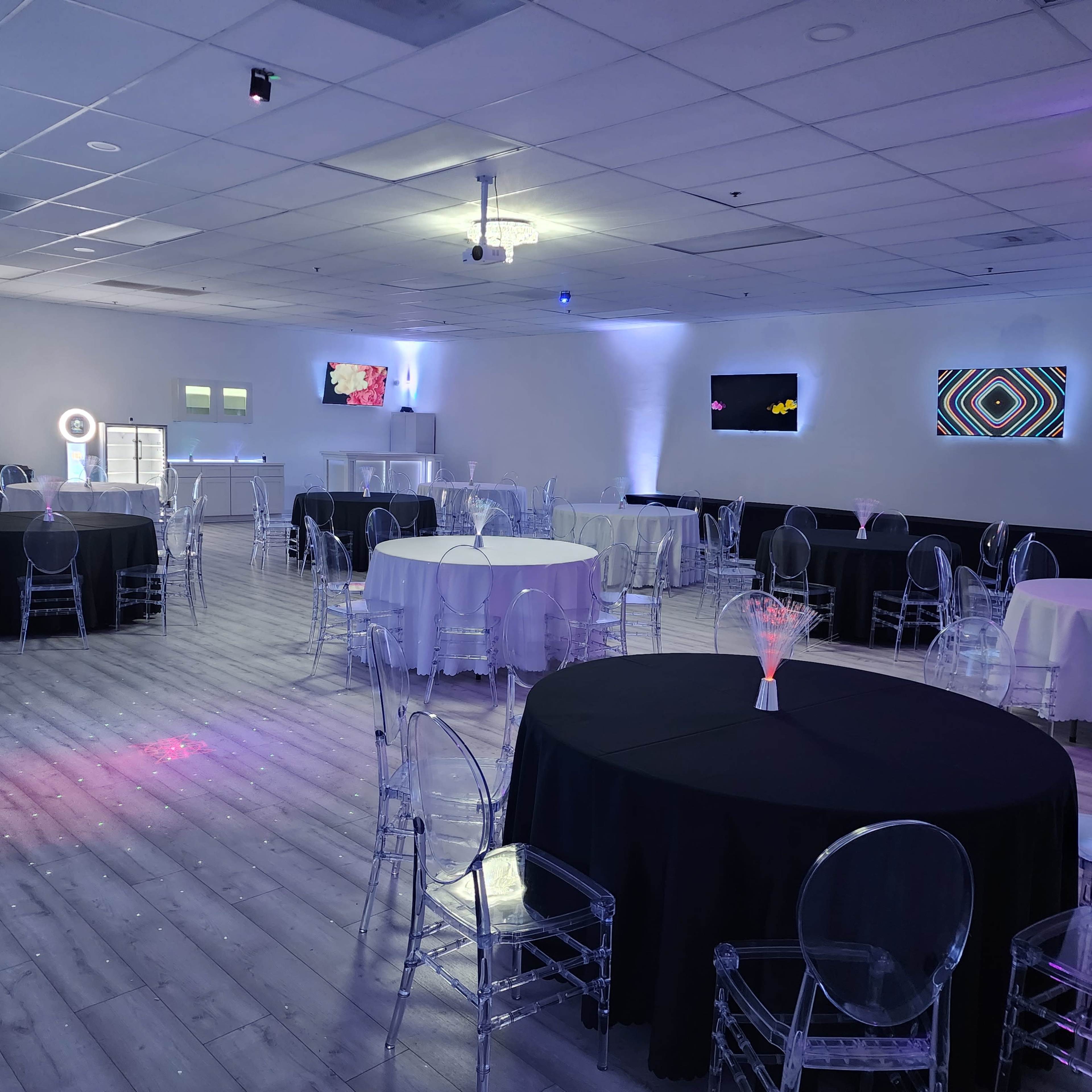 A spacious event room with several round tables, some covered in white and others in black tablecloths, surrounded by transparent chairs and illuminated by colored lights.