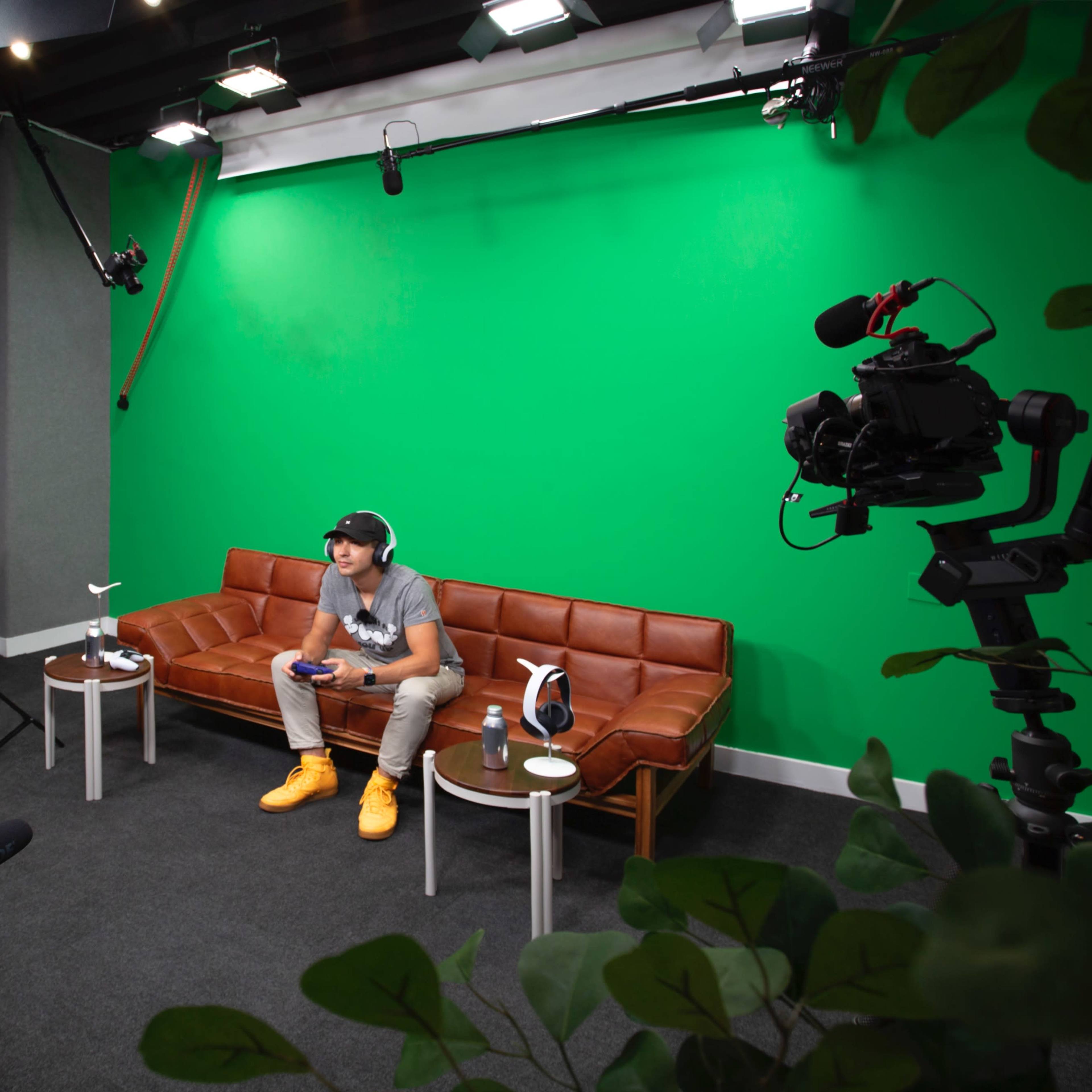 A young man sits on a brown leather couch in a studio with a green screen background, holding a video game controller, surrounded by cameras and lighting equipment.