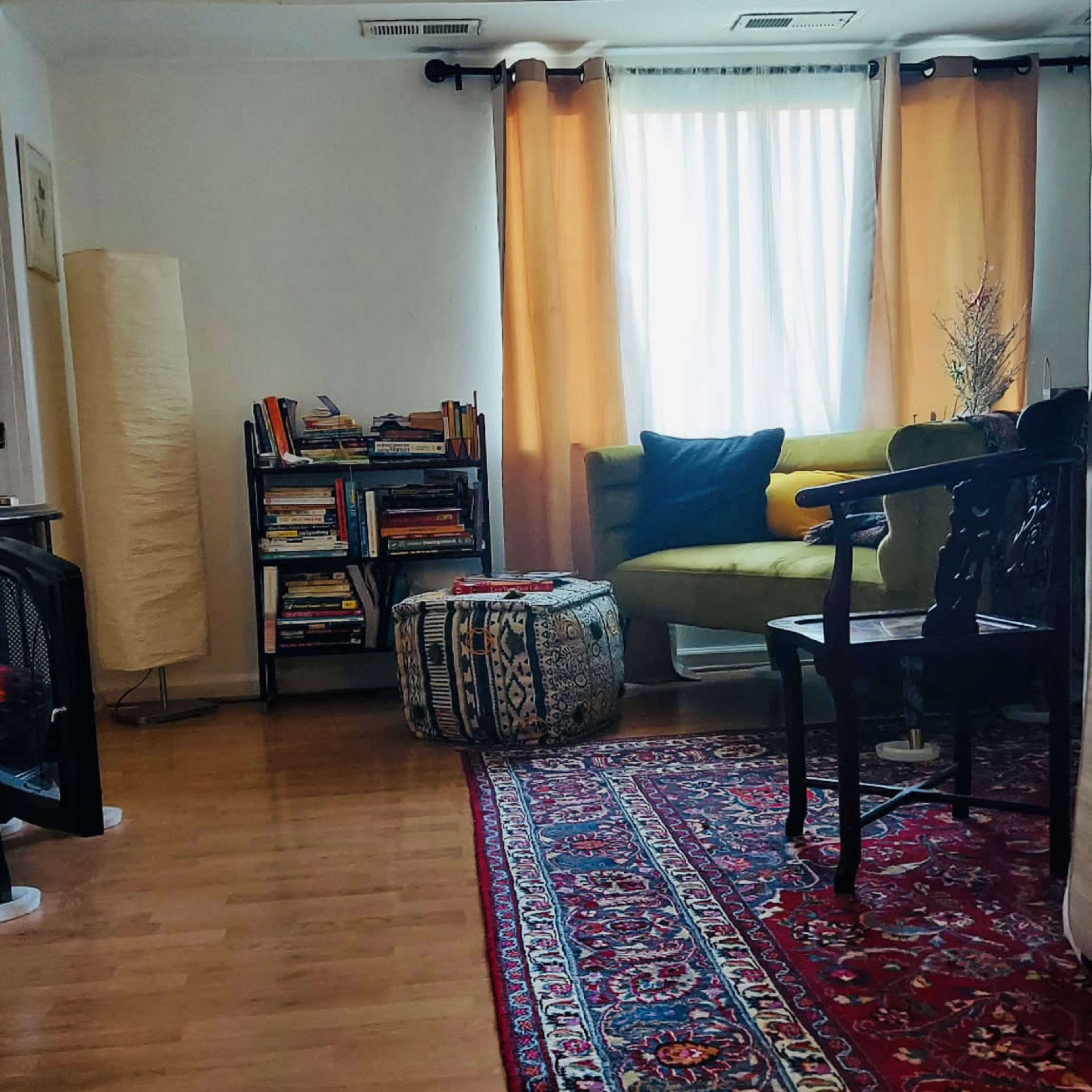 A well-lit living room features a green sofa, a bookshelf filled with books, a patterned ottoman, and a decorative rug on the wooden floor.