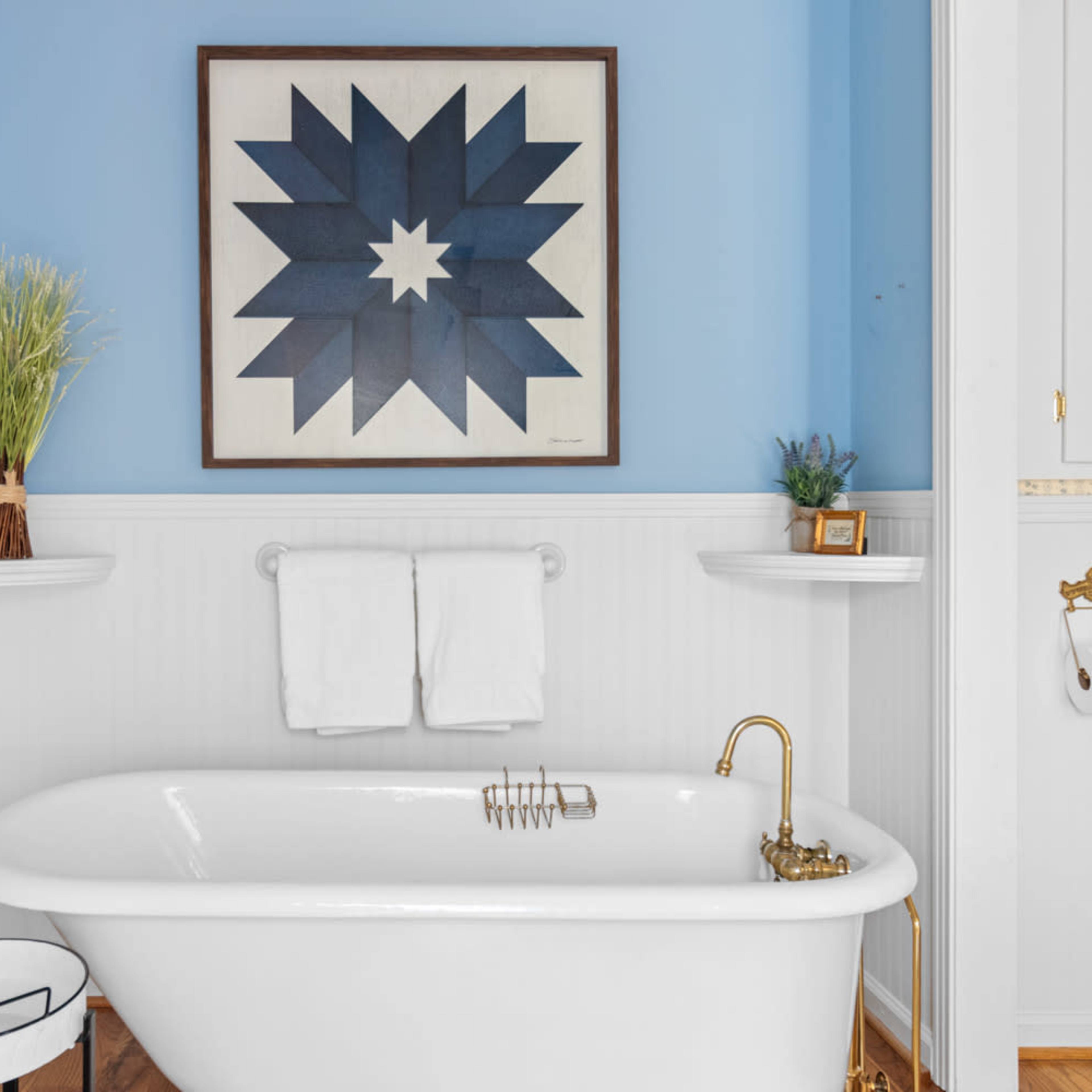 A freestanding bathtub is positioned against a light blue wall, accented by two small shelves with plants and a geometric artwork above.
