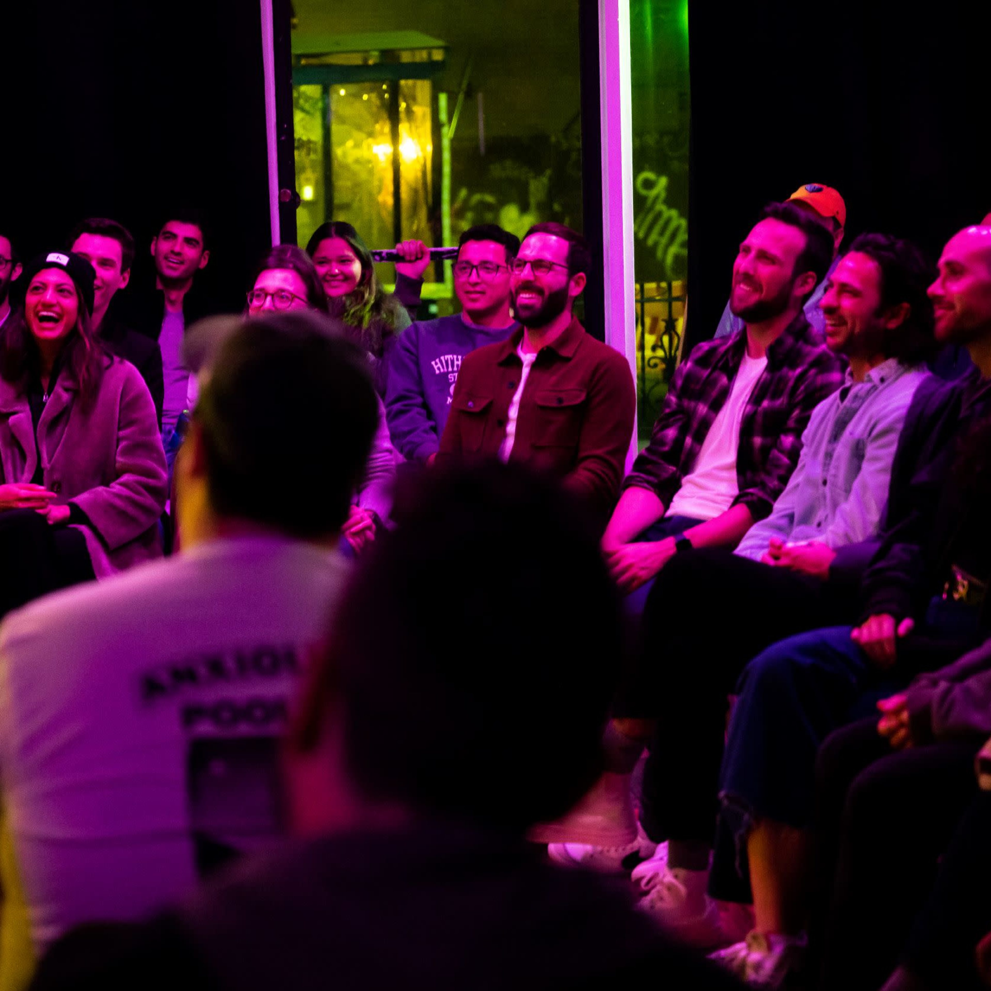 A group of about twenty people sits in a dimly lit room, facing a performer on stage, with colorful lighting creating a vibrant atmosphere.