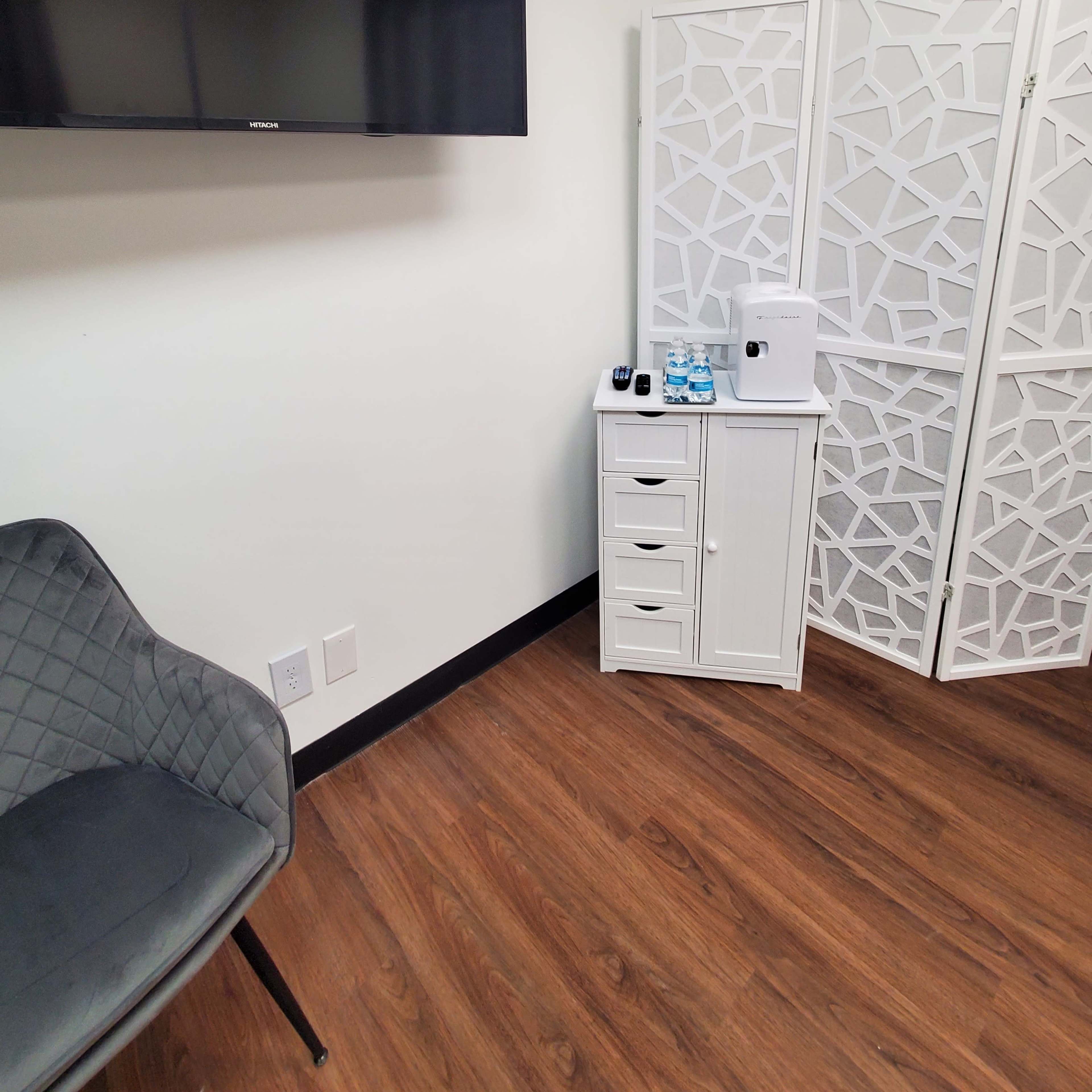 A small room with a TV mounted on the wall, a decorative partition, a white storage cabinet, and a gray chair on a wooden floor.