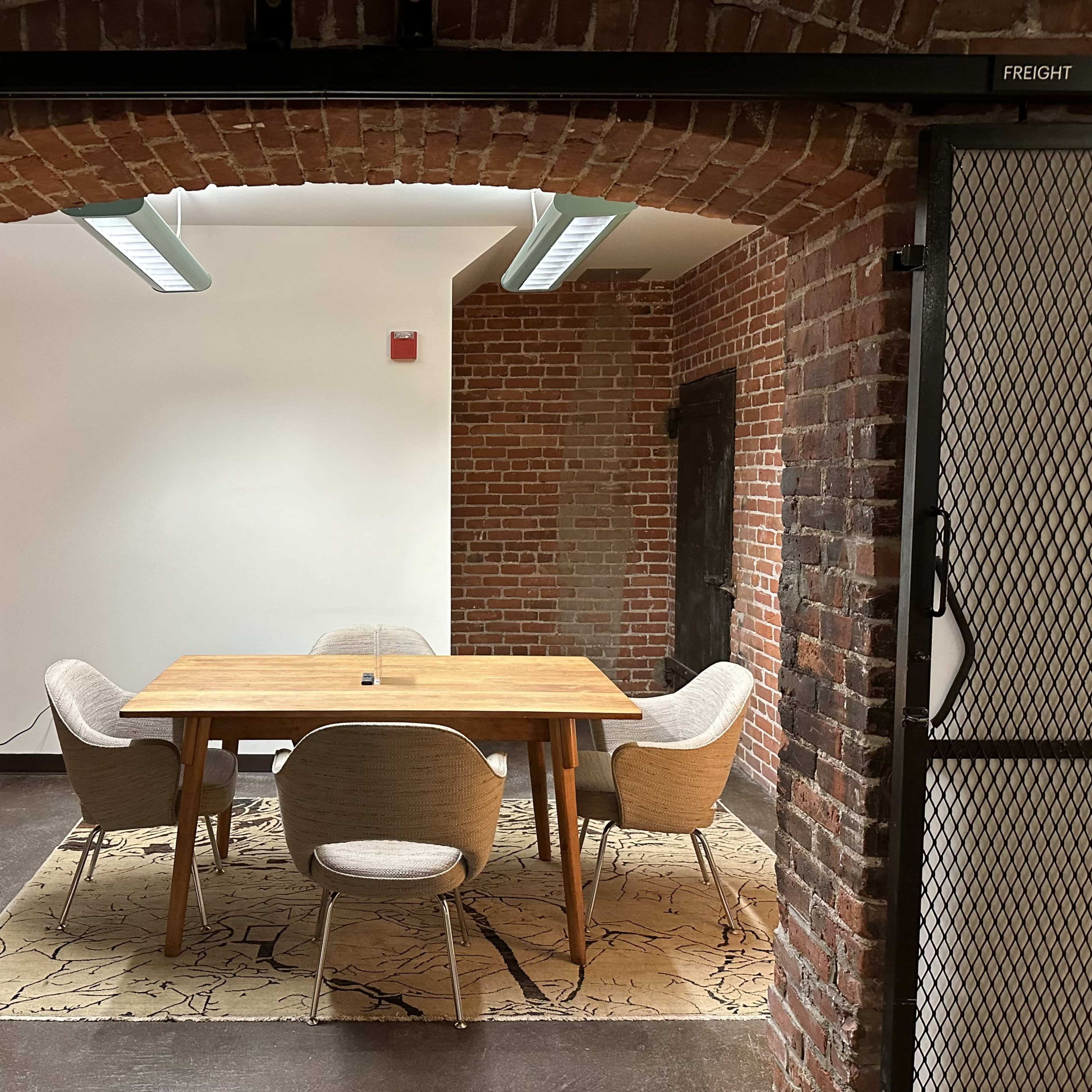 A brick-walled meeting room features a wooden table surrounded by four upholstered chairs on a patterned rug.