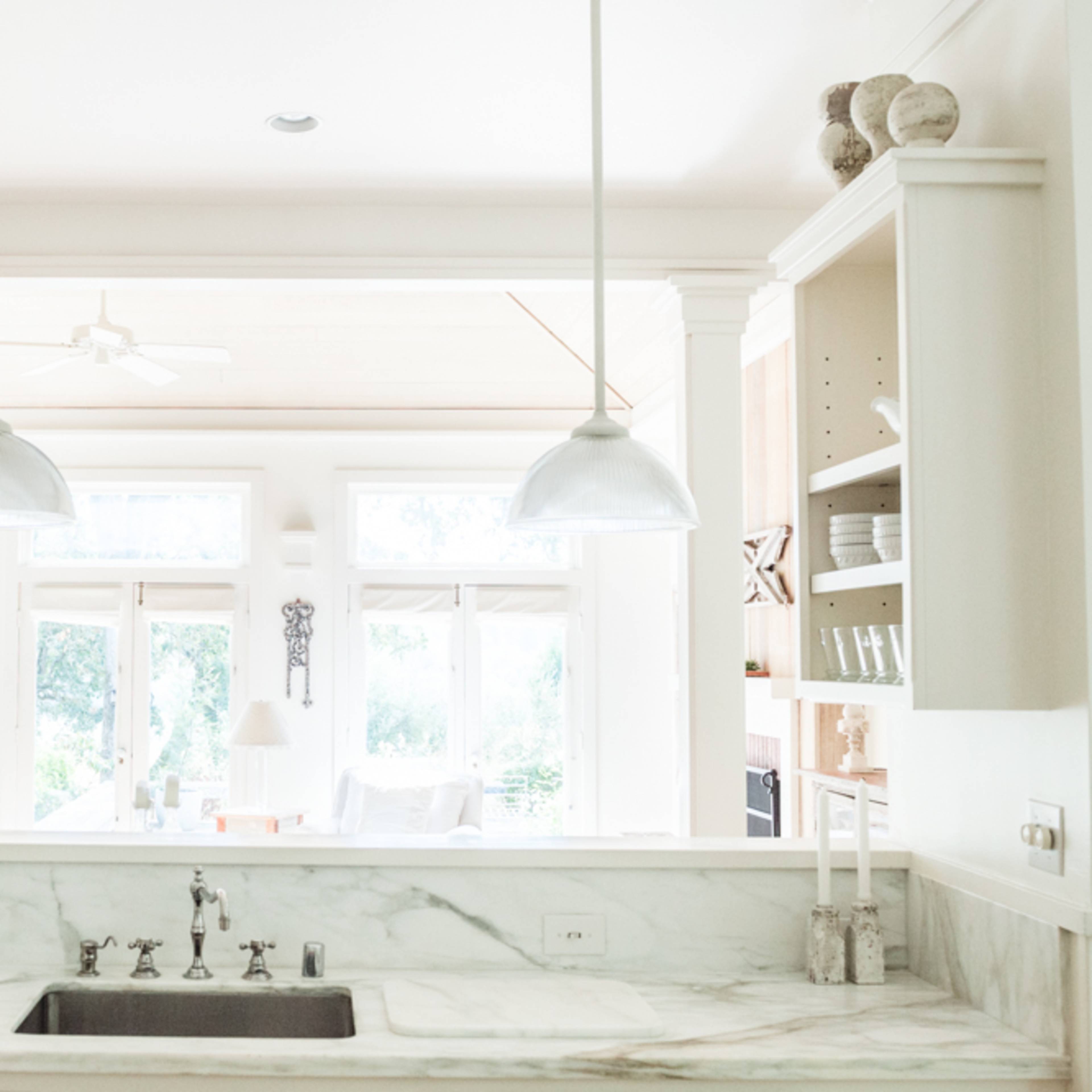 A bright kitchen with marble countertops, open shelving displaying glassware, and a view of a living area through large windows.