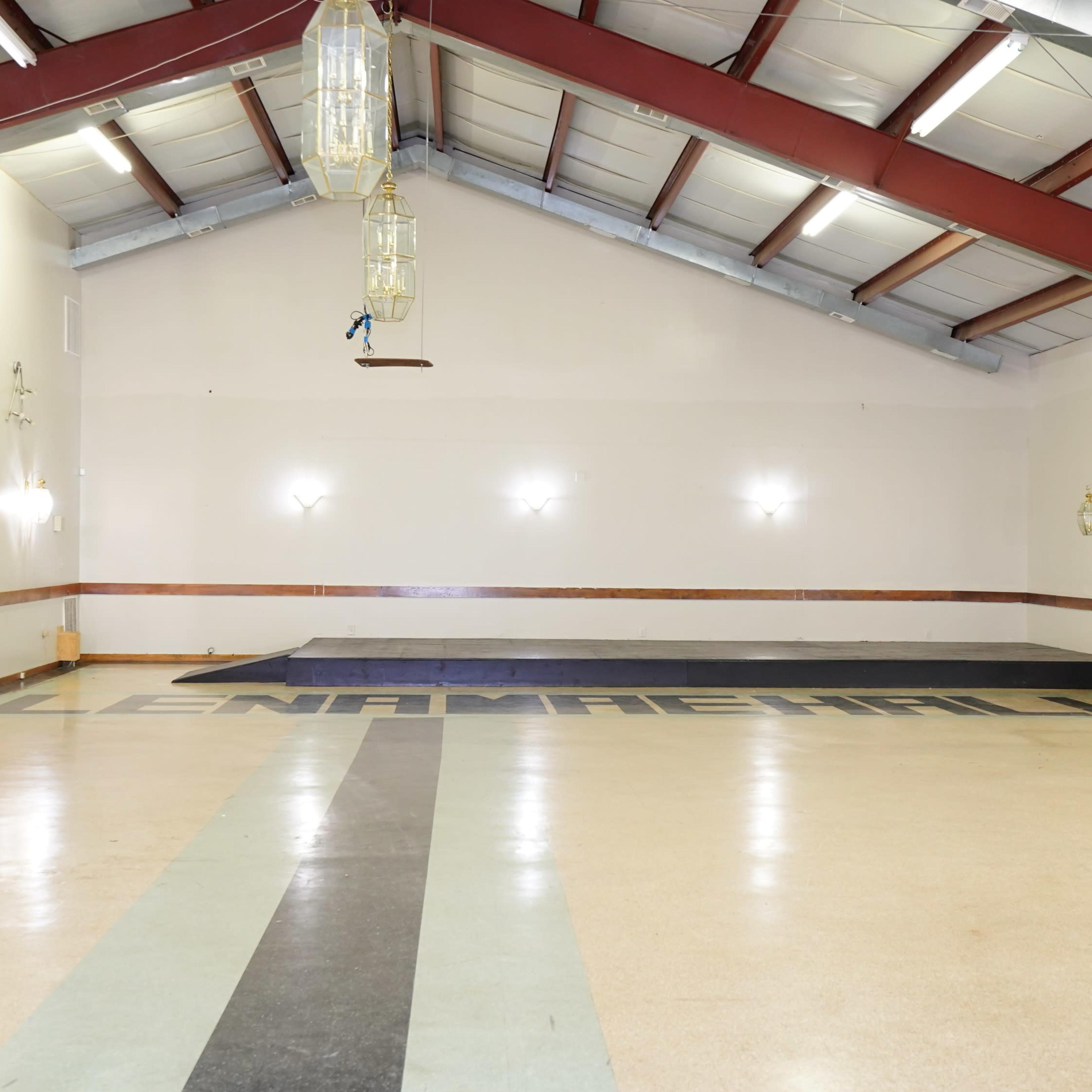 An empty hall with a wooden platform at one end and "LENAMAREHALL" painted on the floor.