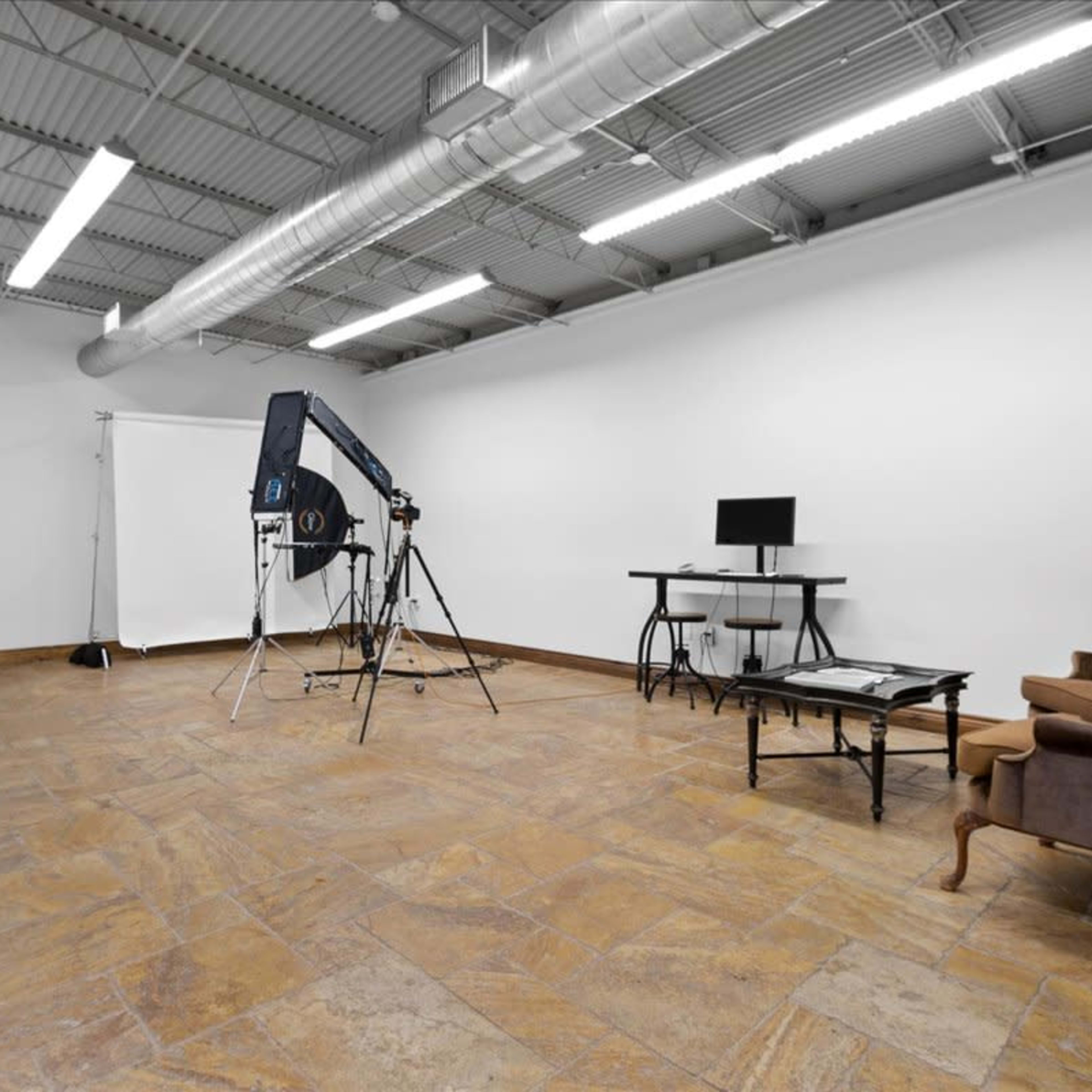 An empty studio space featuring a photography backdrop, a computer setup on a desk, and a large chair against a wall with tiled flooring.