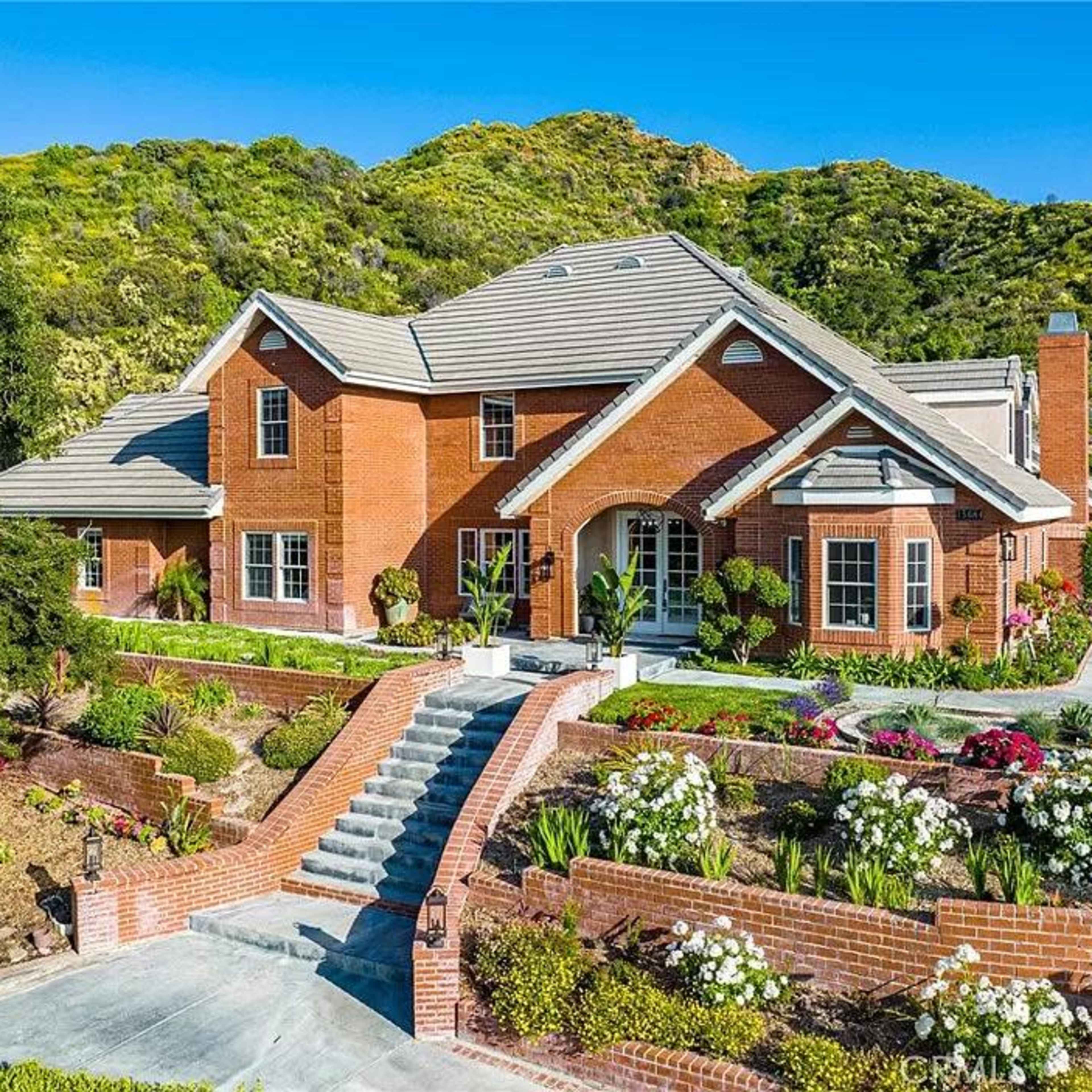 A large brick house with a steep roof is situated on a landscaped property featuring flowers and greenery, against a hilly background.