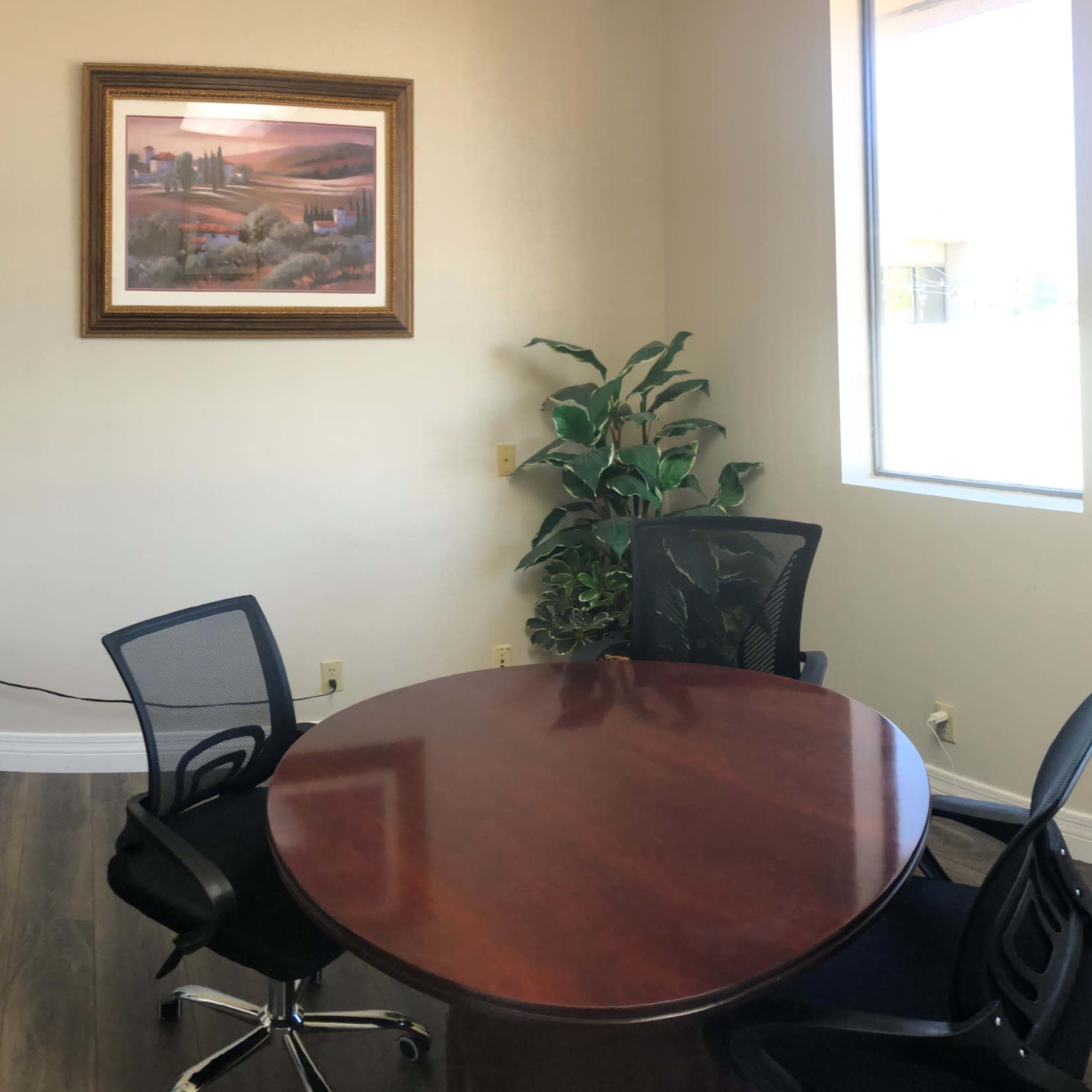 A small conference room featuring a round wooden table surrounded by four black mesh chairs, with a potted plant and a framed painting on the walls.