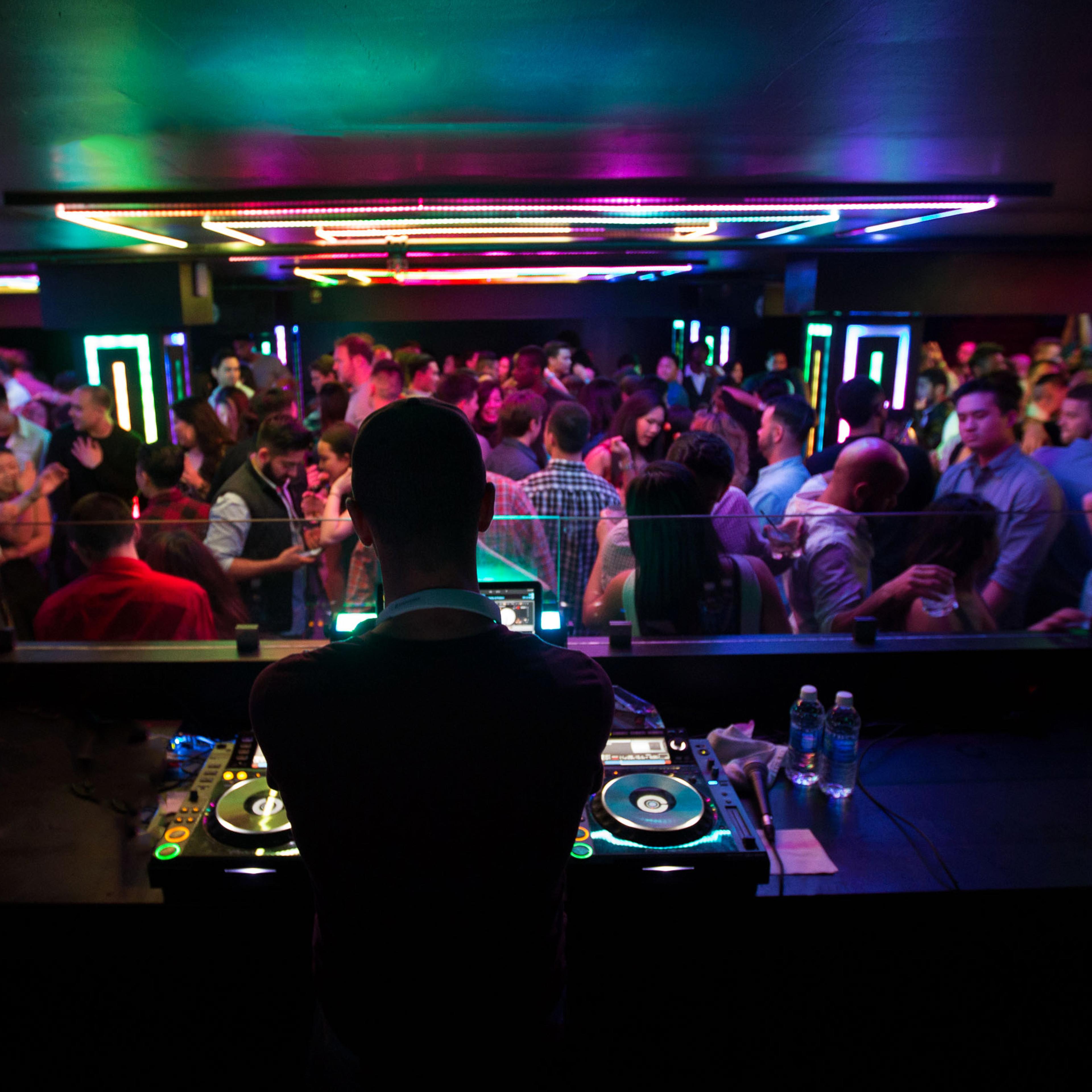 A DJ stands behind a mixing console while a crowd dances in a vibrant, neon-lit nightclub.