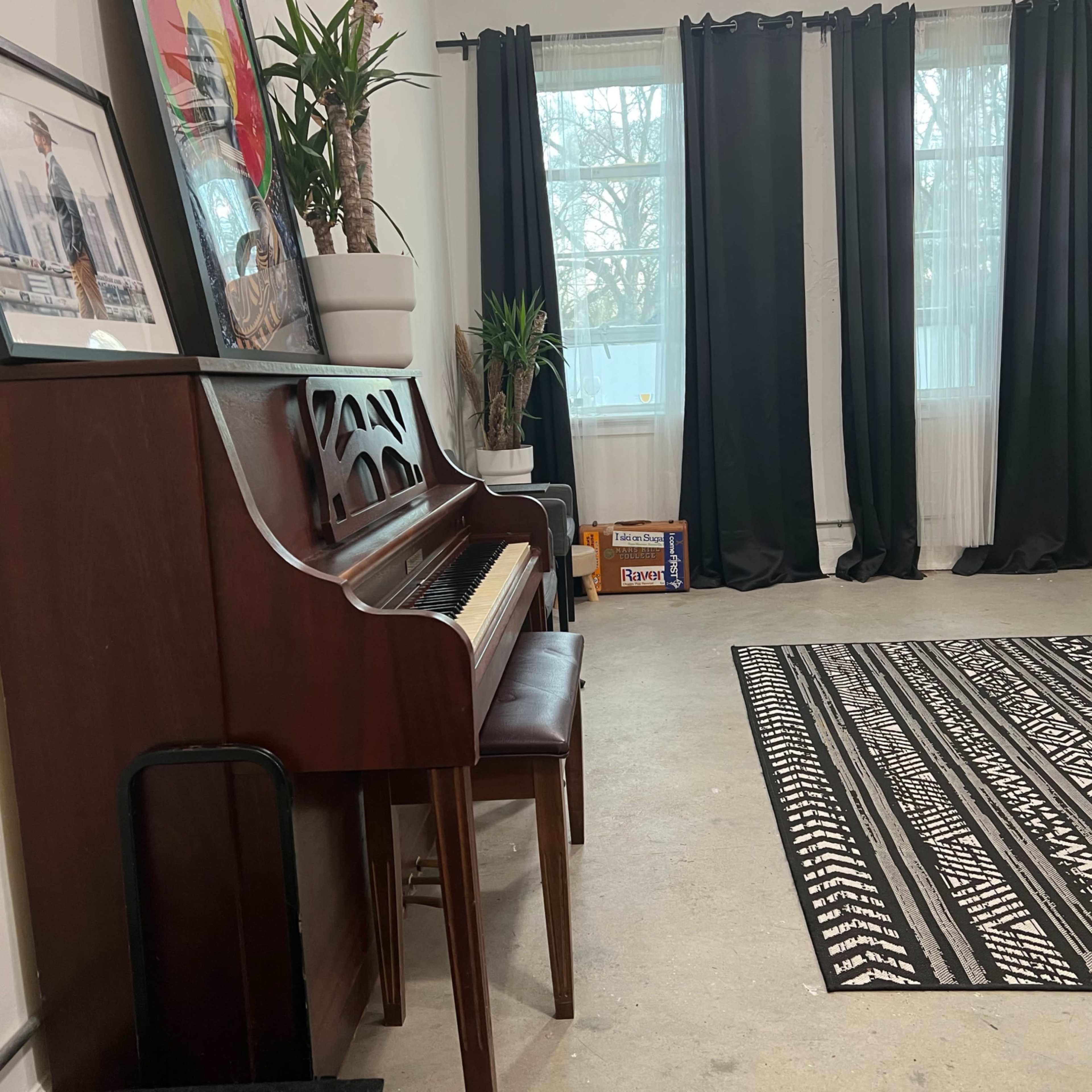 A room with a piano against one wall, a patterned rug on the floor, and large windows covered by dark curtains.