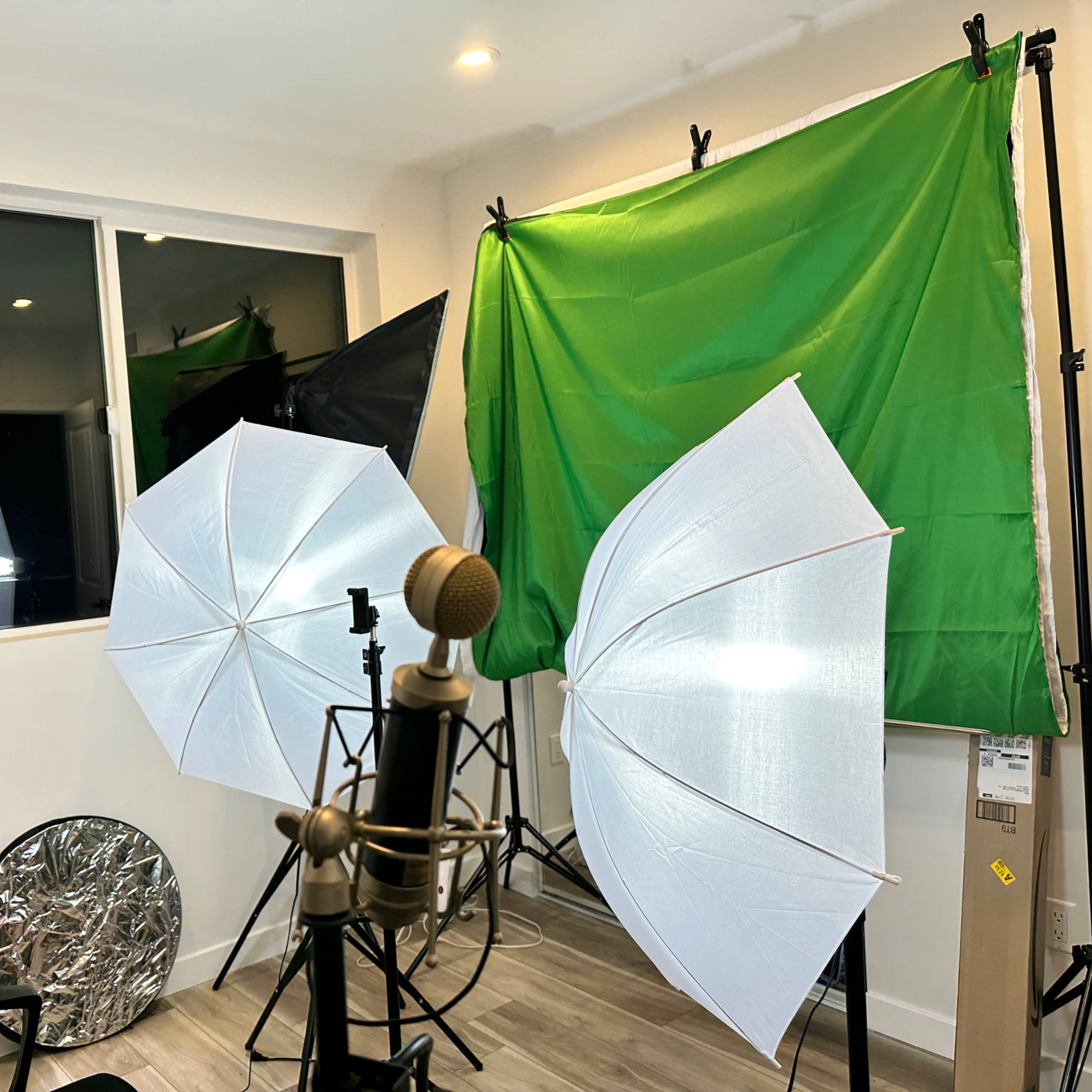 A small studio setup with green and black backdrops, large white umbrellas for lighting, and a microphone in the foreground.