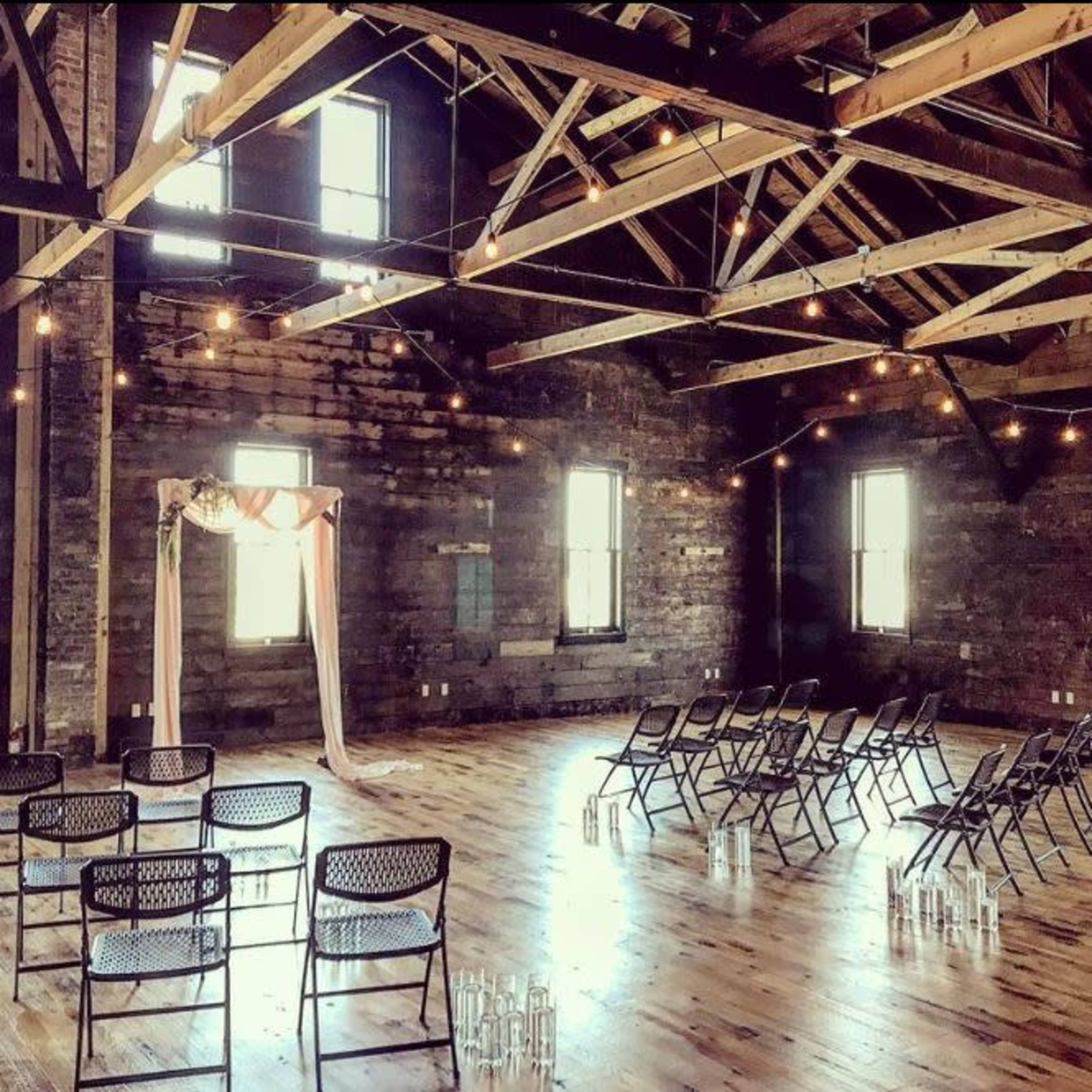 The image shows a spacious, rustic event venue set up with rows of chairs facing a decorative arch, illuminated by hanging string lights.
