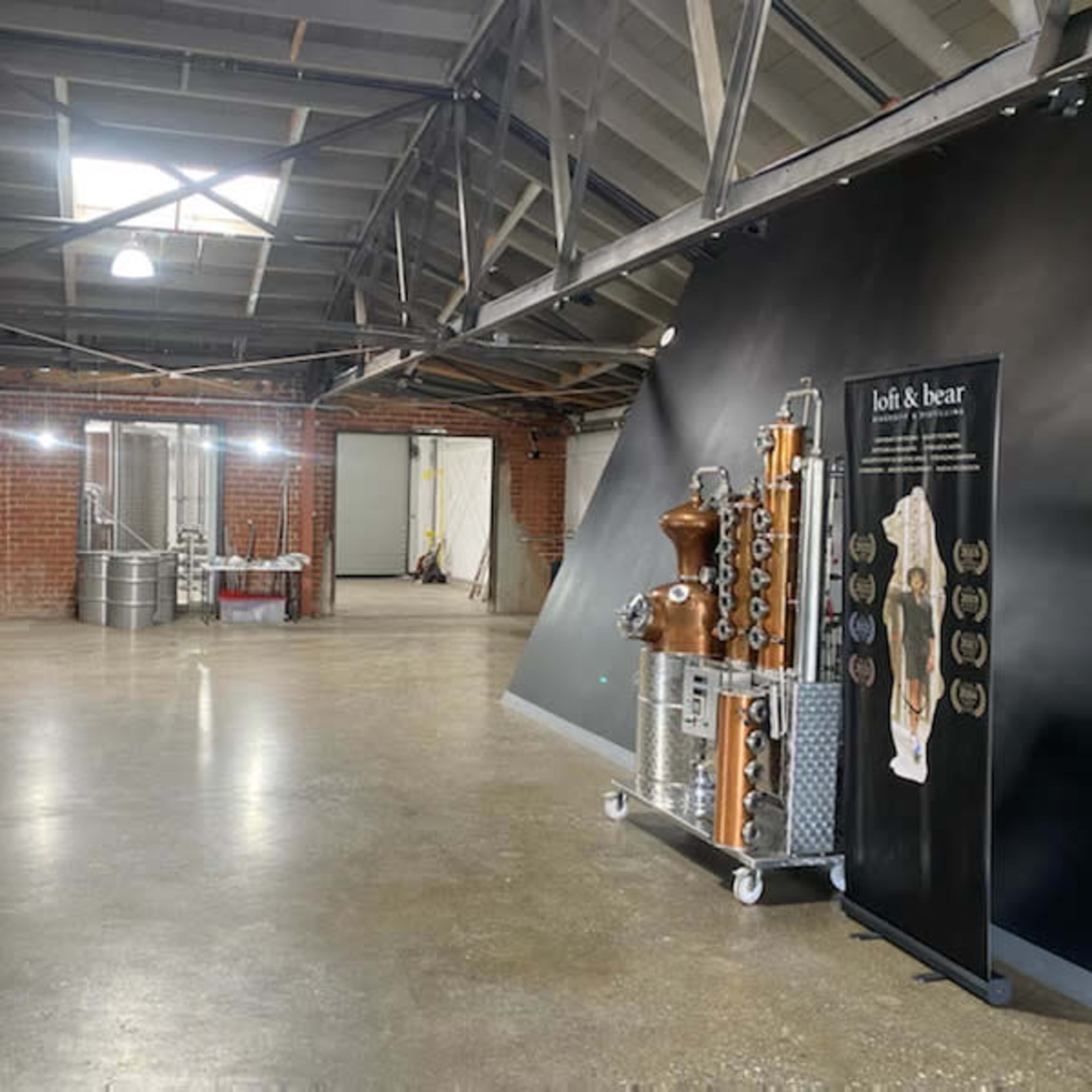 An industrial space with polished concrete floors, exposed beams, and a display featuring distillation equipment alongside large metal tanks in the background.
