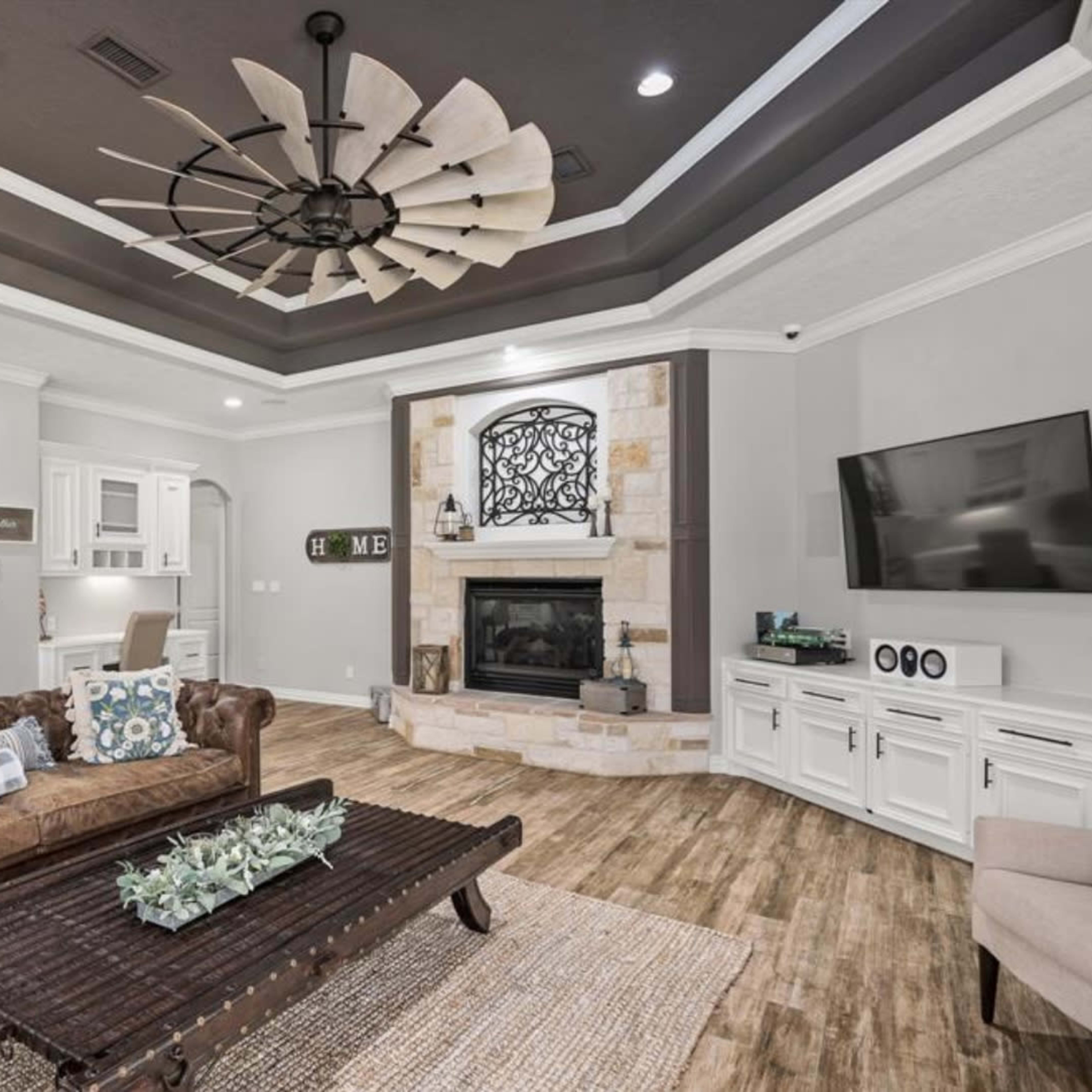 A living room featuring a brown leather sofa, a wooden coffee table, a stone fireplace with a television above it, and a ceiling fan.