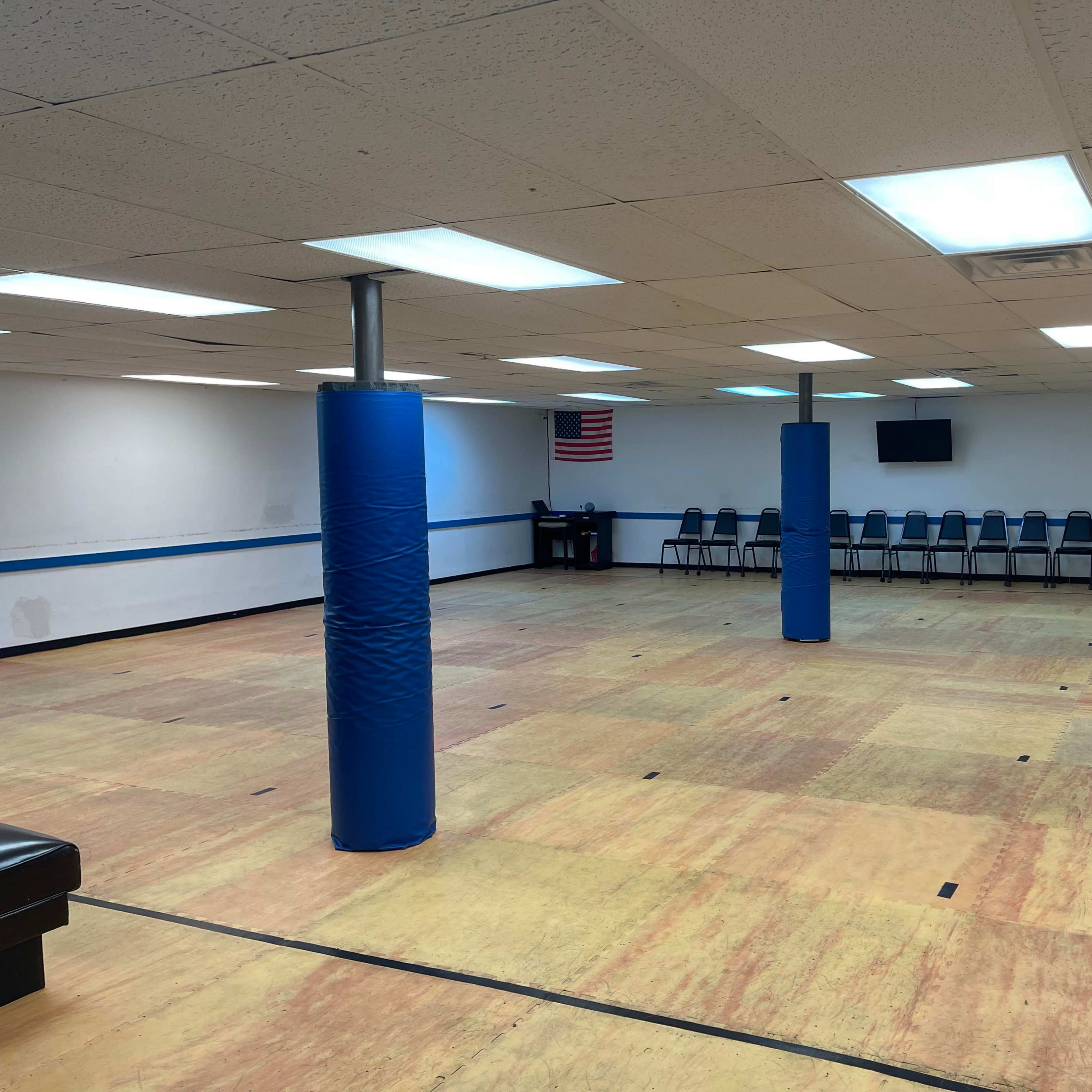 An empty room with a wooden floor, blue padded columns, and rows of chairs along the walls.