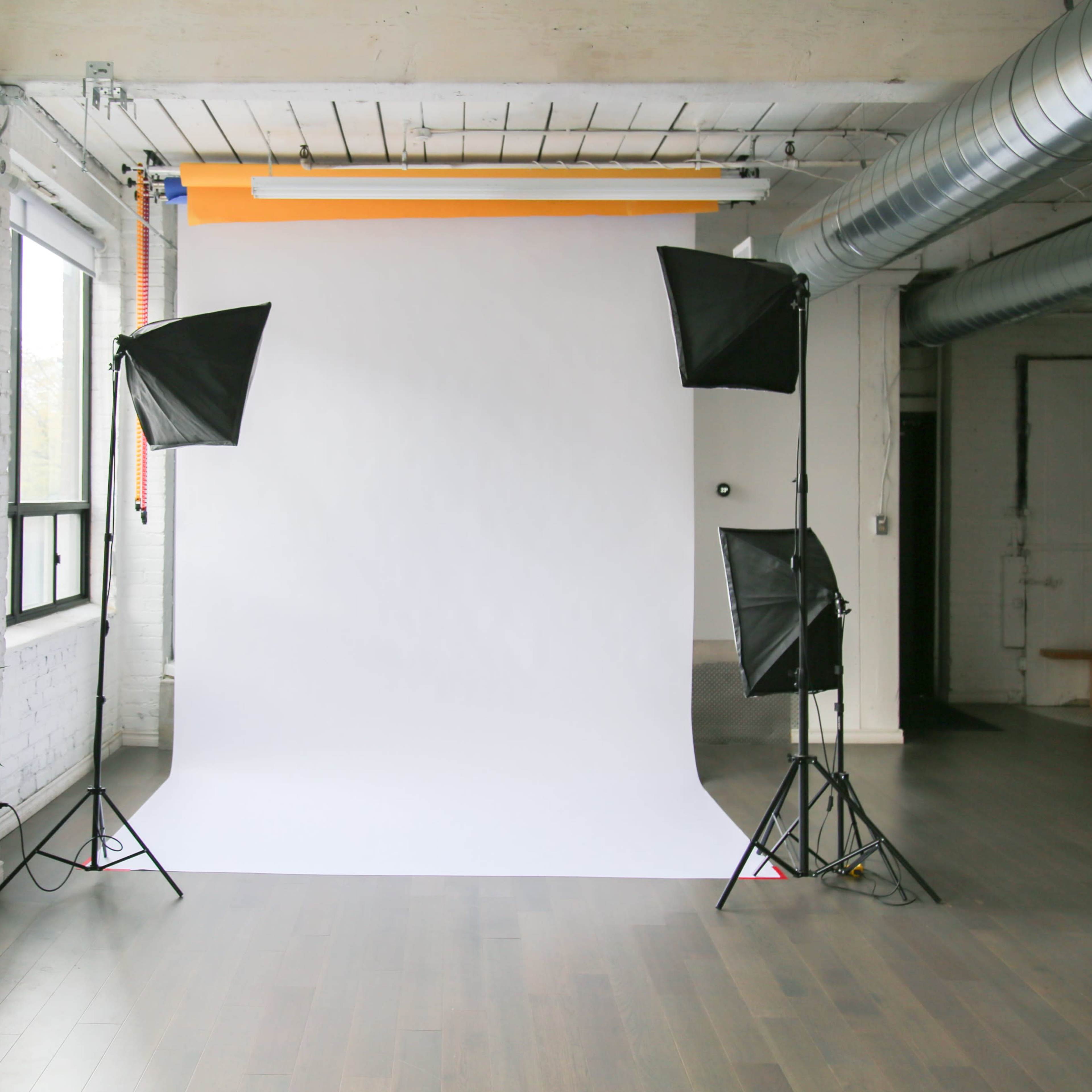 A photography studio features a white backdrop with two softbox lights on either side, and a large plant in the corner.