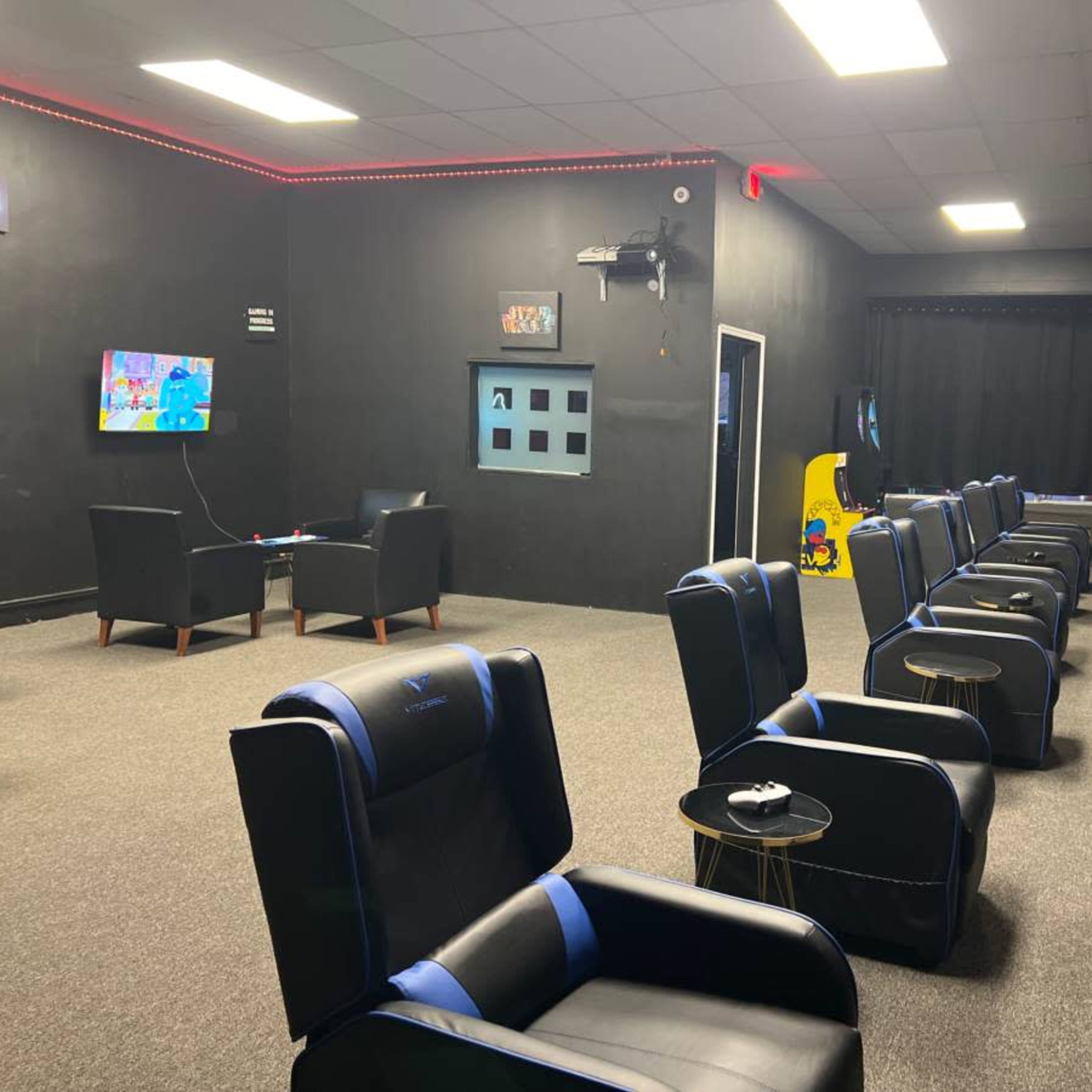 A gaming lounge with several black and blue chairs arranged in rows facing wall-mounted televisions, along with arcade games at one end of the room.