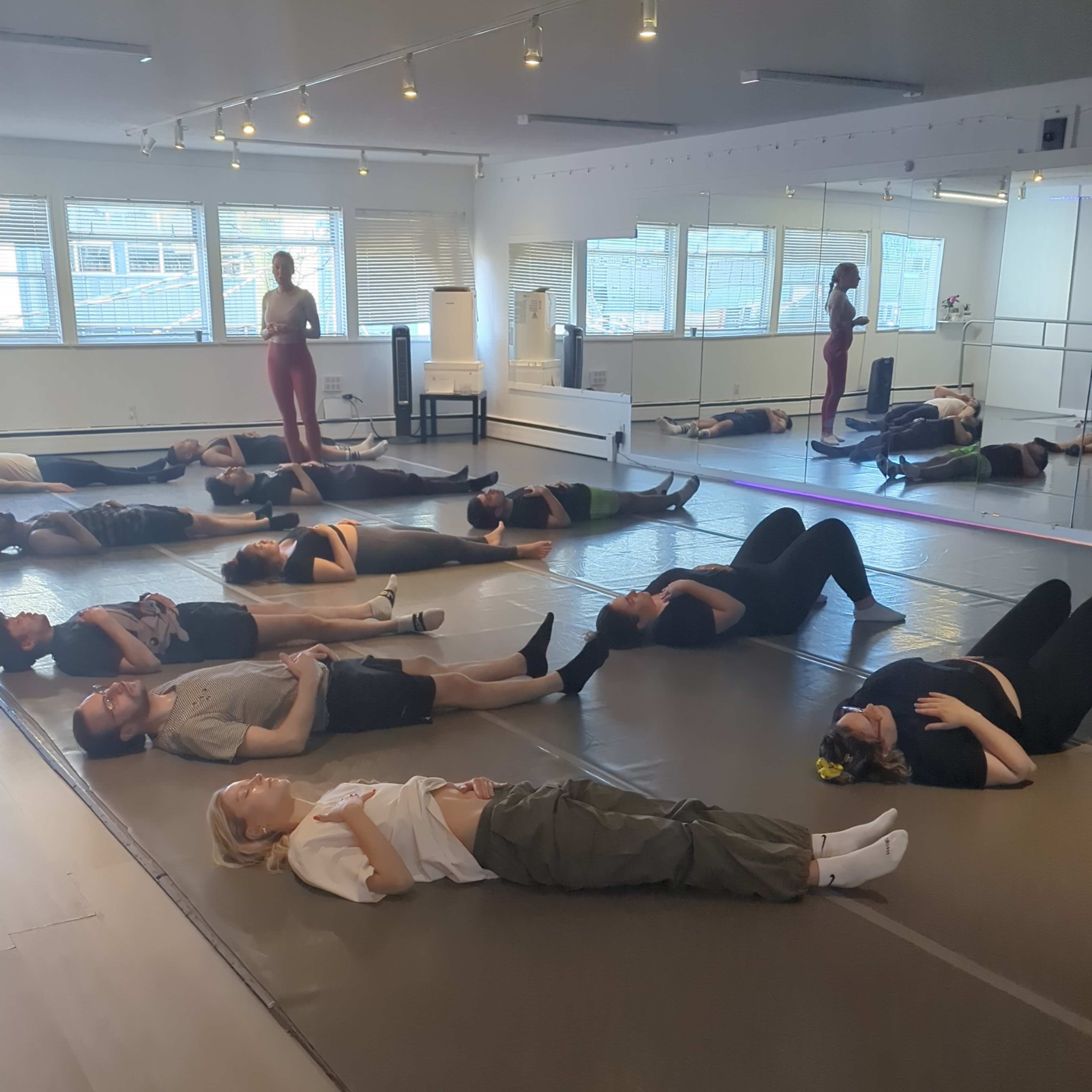 A group of participants is lying on mats in a spacious studio, engaged in a floor exercise while others stand nearby.