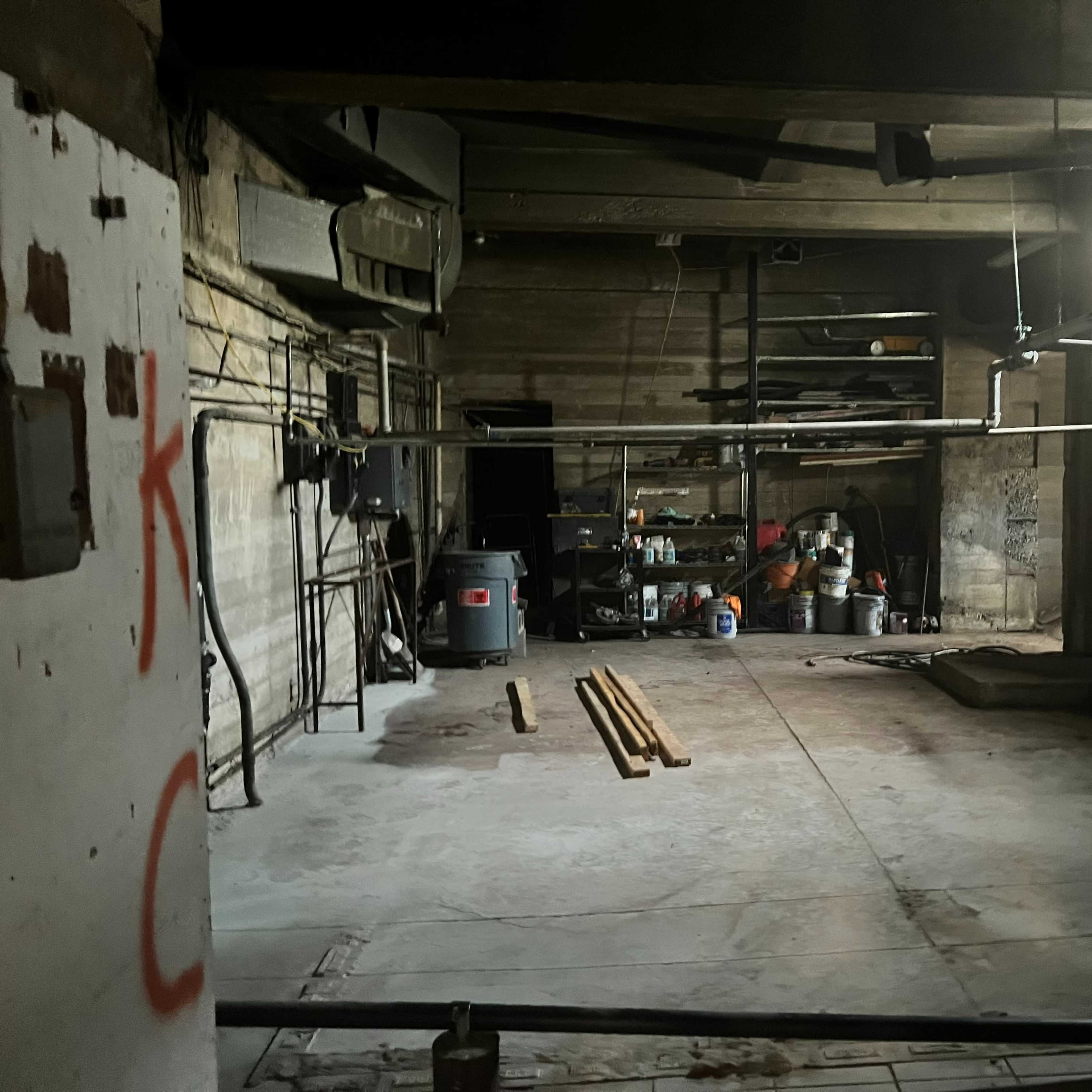 A dimly lit, empty basement space with exposed wooden beams and various tools and containers stacked against the walls.