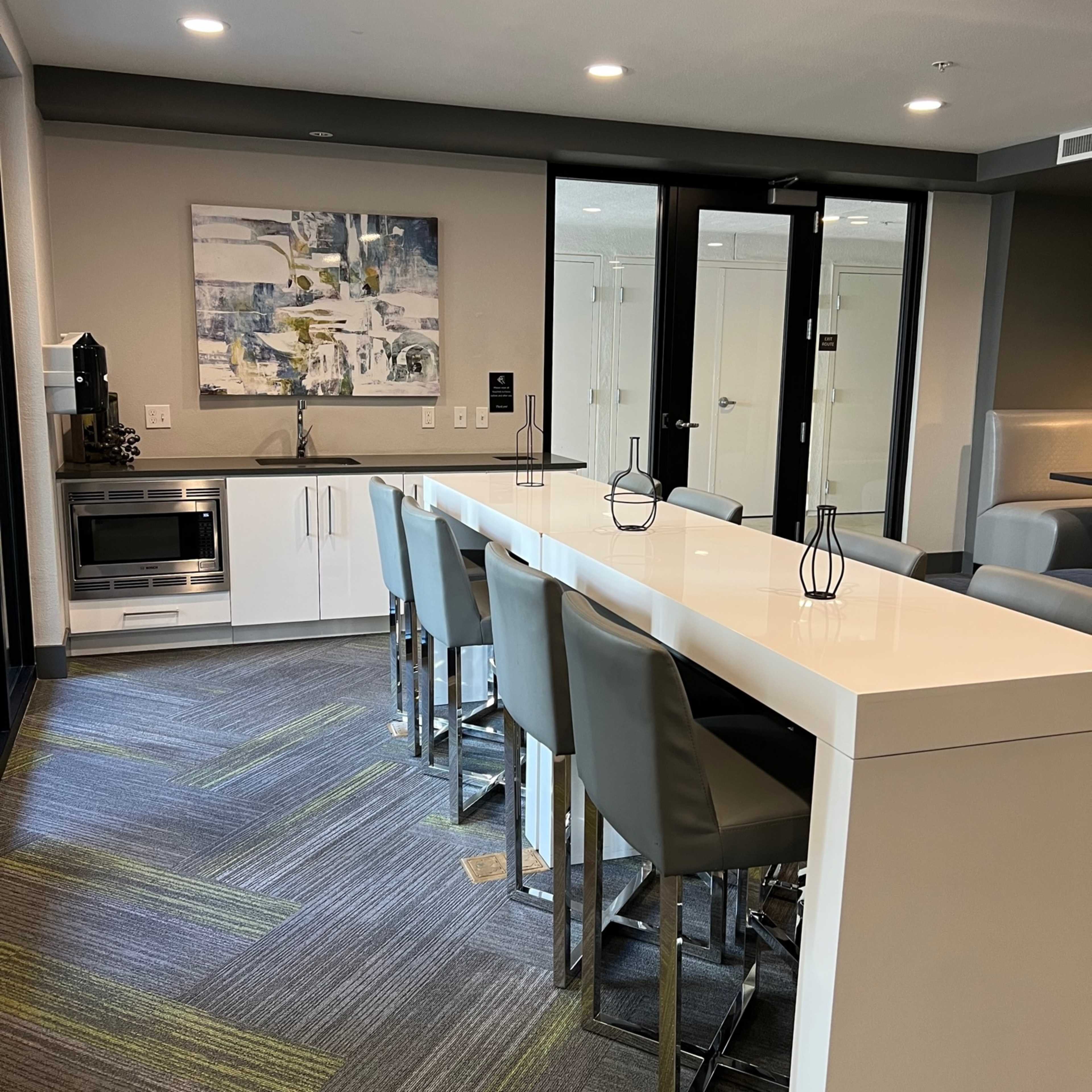A modern kitchen area with a long white table surrounded by gray high-backed chairs, a microwave, and a small kitchenette on the side.