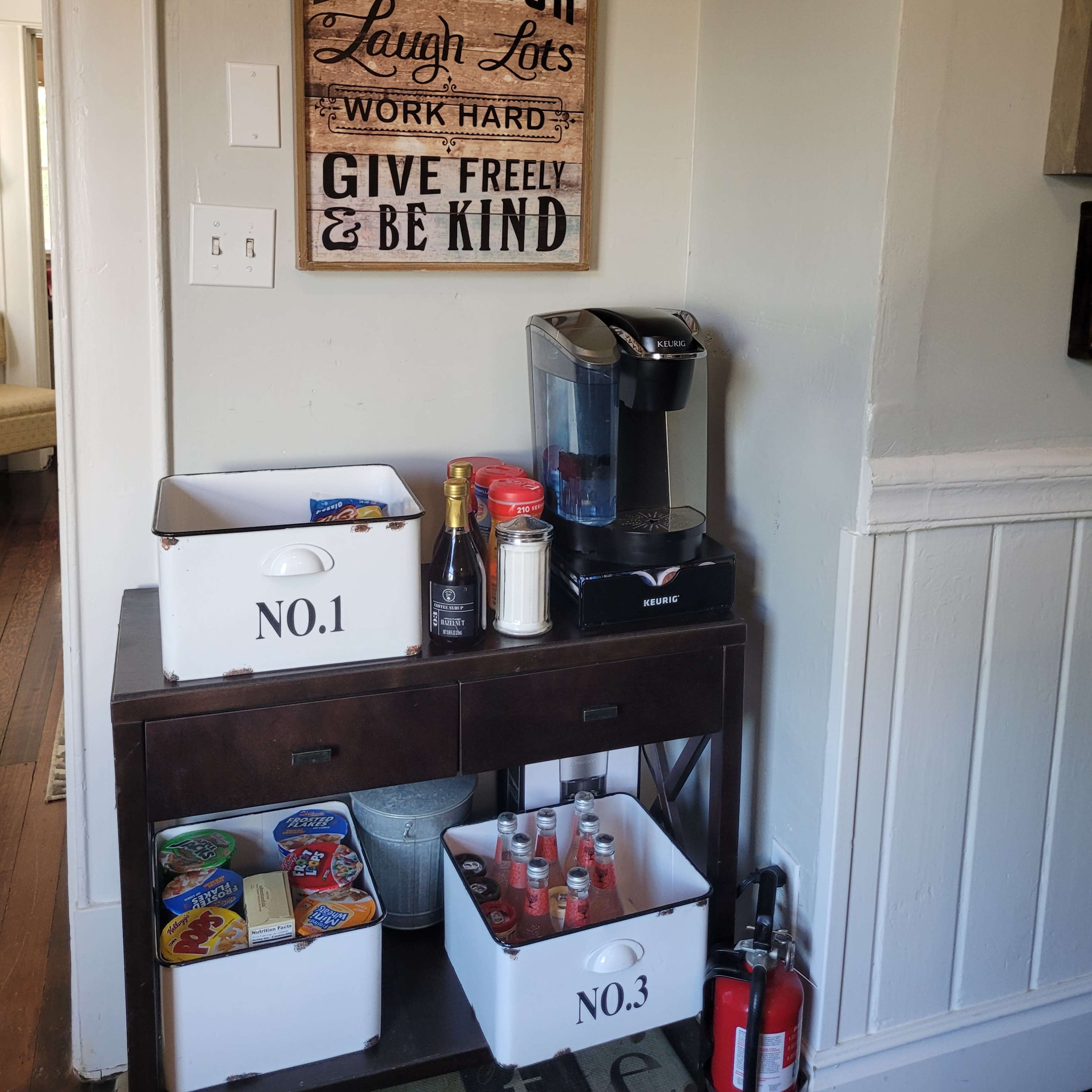 A corner of a room with a coffee maker on a small table, flanked by storage boxes labeled "NO. 1" and "NO. 3," and a decorative sign with encouraging phrases on the wall.
