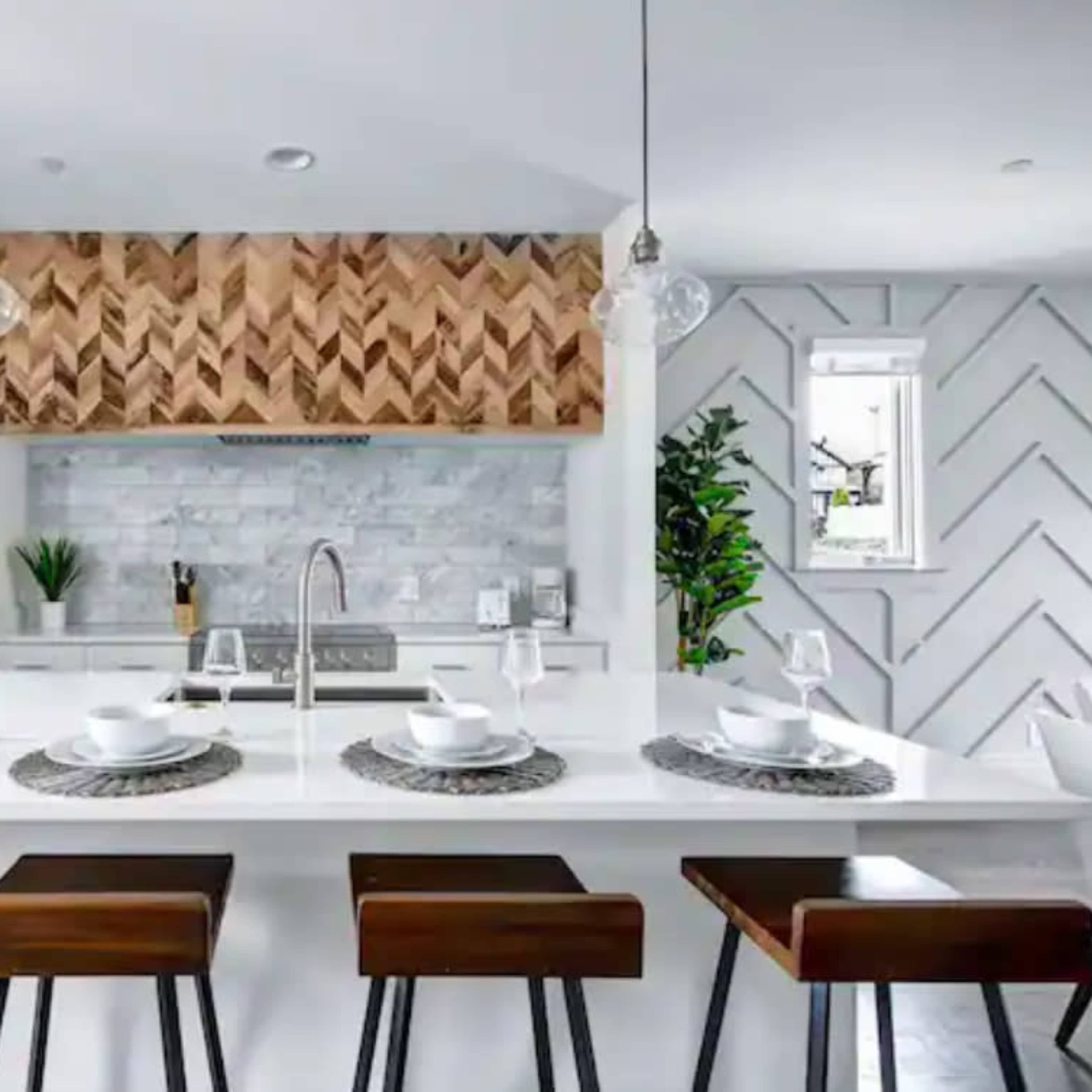 A modern kitchen features a white island with four wooden stools, a herringbone wood accent above the stove, and stone backsplash.